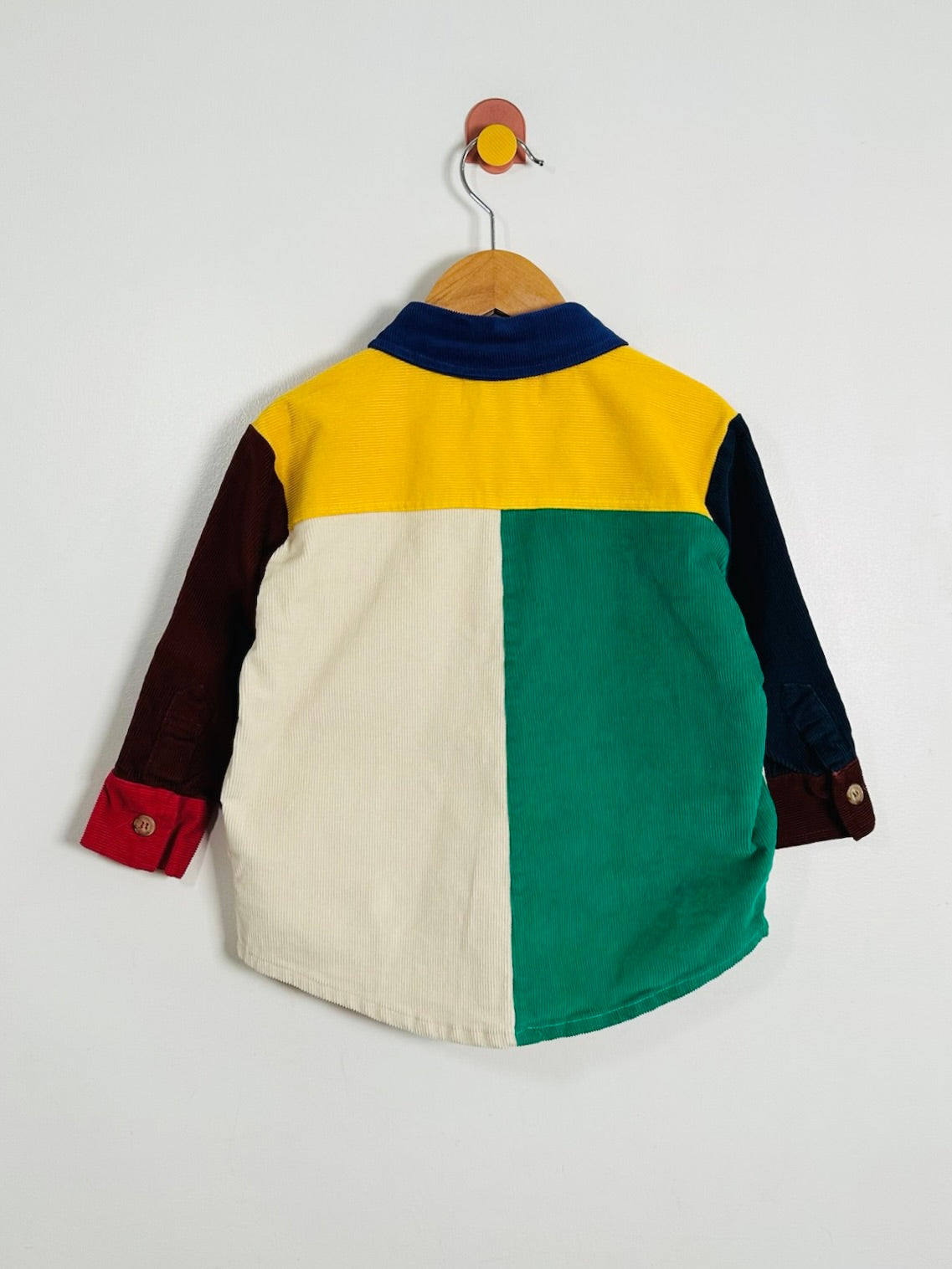 A color-blocked long-sleeve shirt for kids, shown from the back, hanging on a wooden hanger against a plain light gray wall. The shirt features a yellow yoke, a cream-colored lower back panel, a green lower back panel, dark navy sleeves, and dark red cuffs.