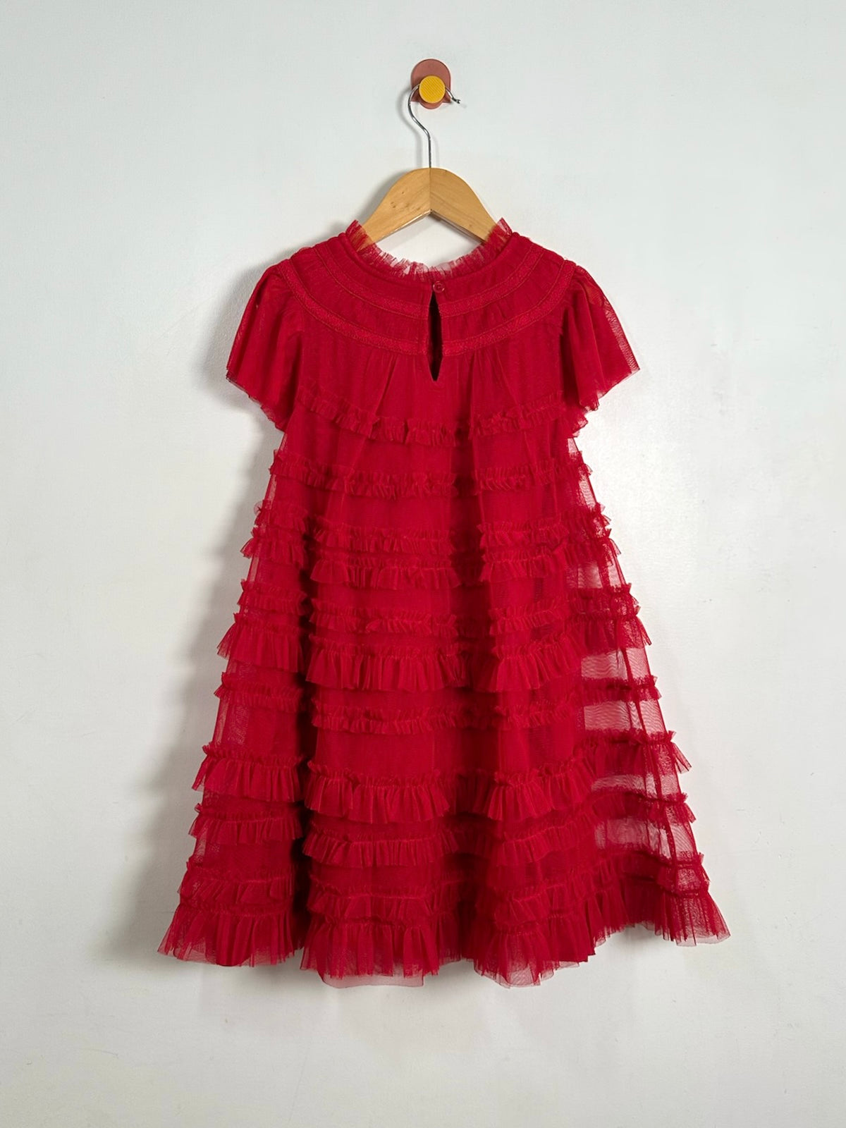 A red tiered tulle dress with short sleeves and a high neckline, hanging on a wooden hanger against a plain white wall.