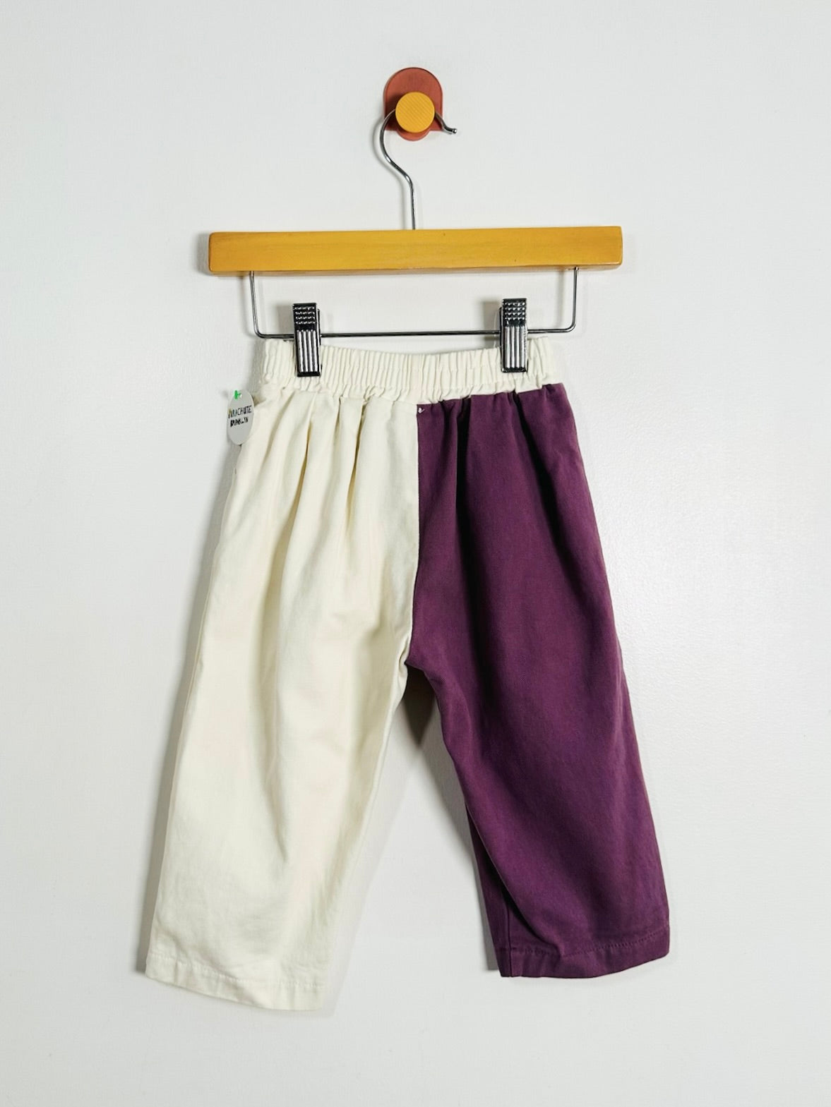A pair of cropped pants hanging from a wooden hanger against a plain white wall. The pants are split down the middle with one side in off-white/cream and the other side in deep purple.