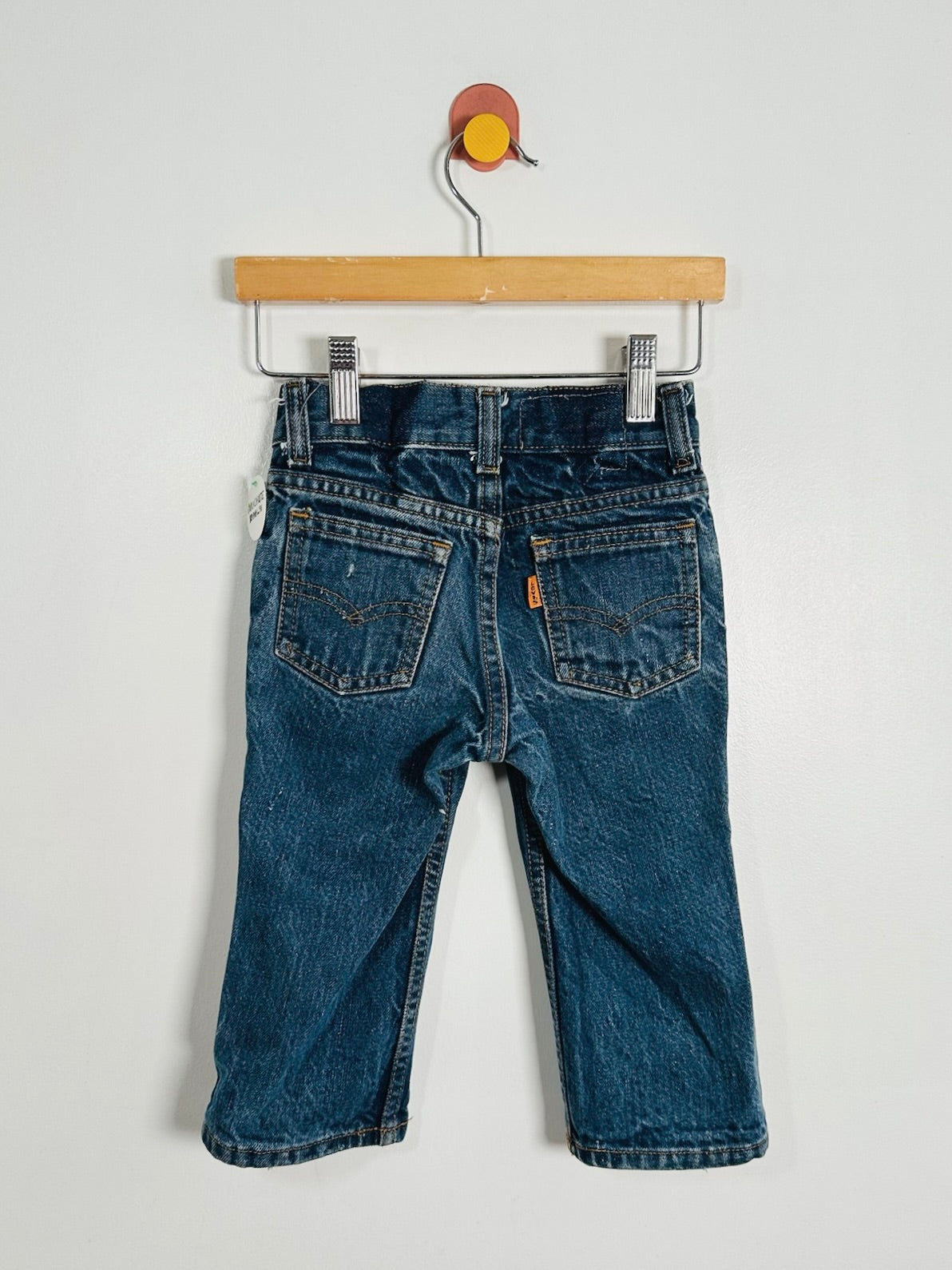 Back view of a pair of blue denim jeans hanging from a wooden hanger against a plain light gray wall.
