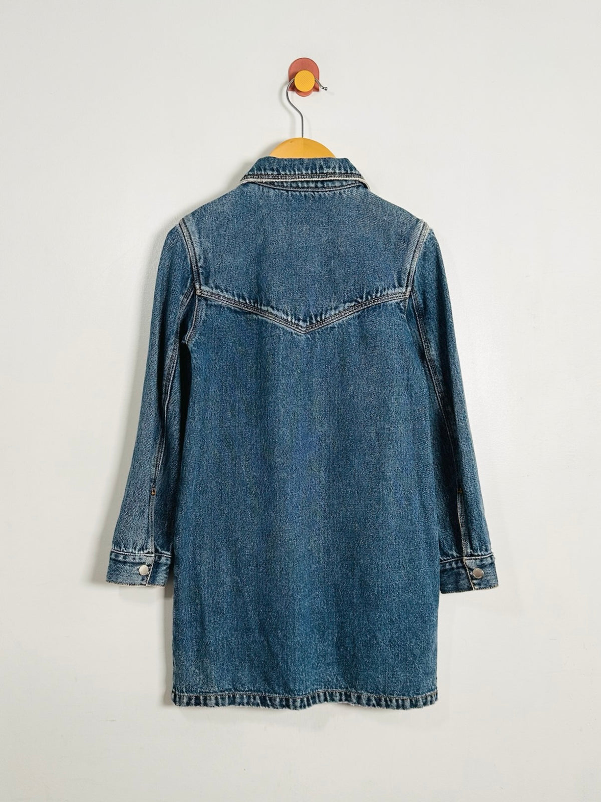 A blue denim shirt jacket shown from the back, hanging on a wooden hanger with a red and yellow hook against a plain off-white wall.