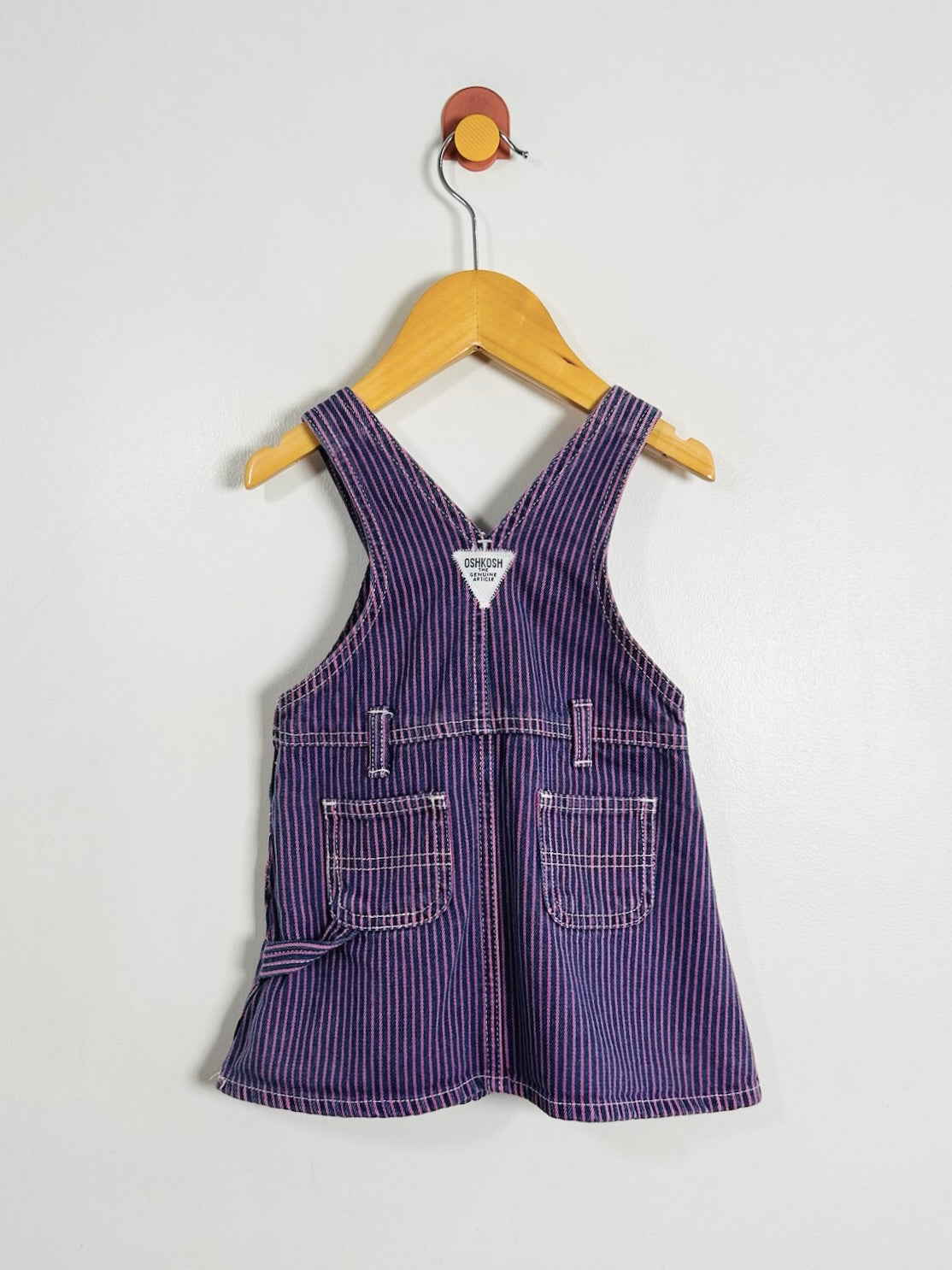 A purple and white striped baby overall dress hanging on a wooden hanger against a plain light gray wall.