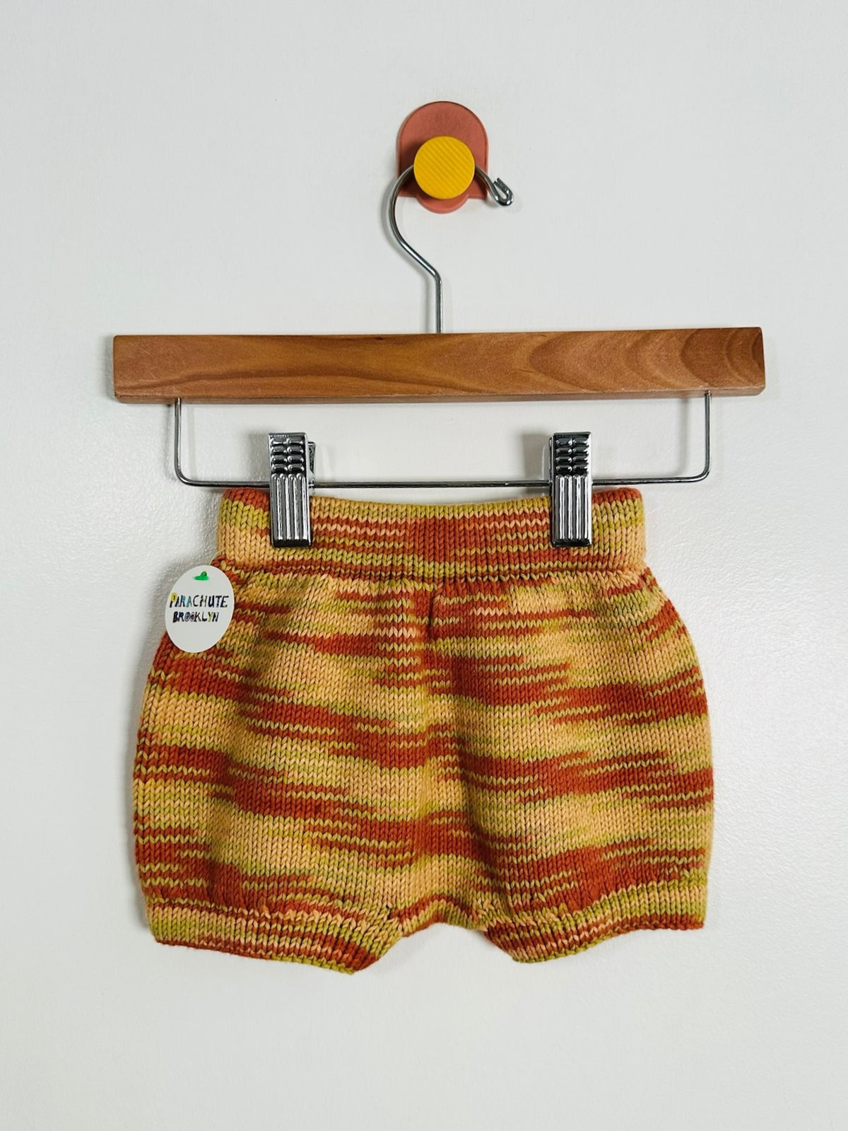 A pair of baby knit shorts with a yellow, orange, and red striped pattern, hanging from a wooden hanger with metal clips against a plain white wall.