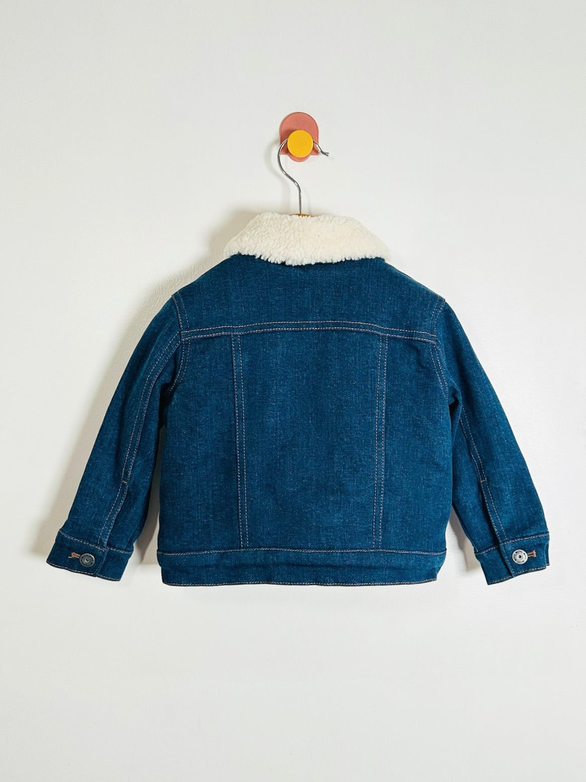 A blue denim jacket with a white sherpa collar, shown from the back on a metal hanger against a plain white wall.