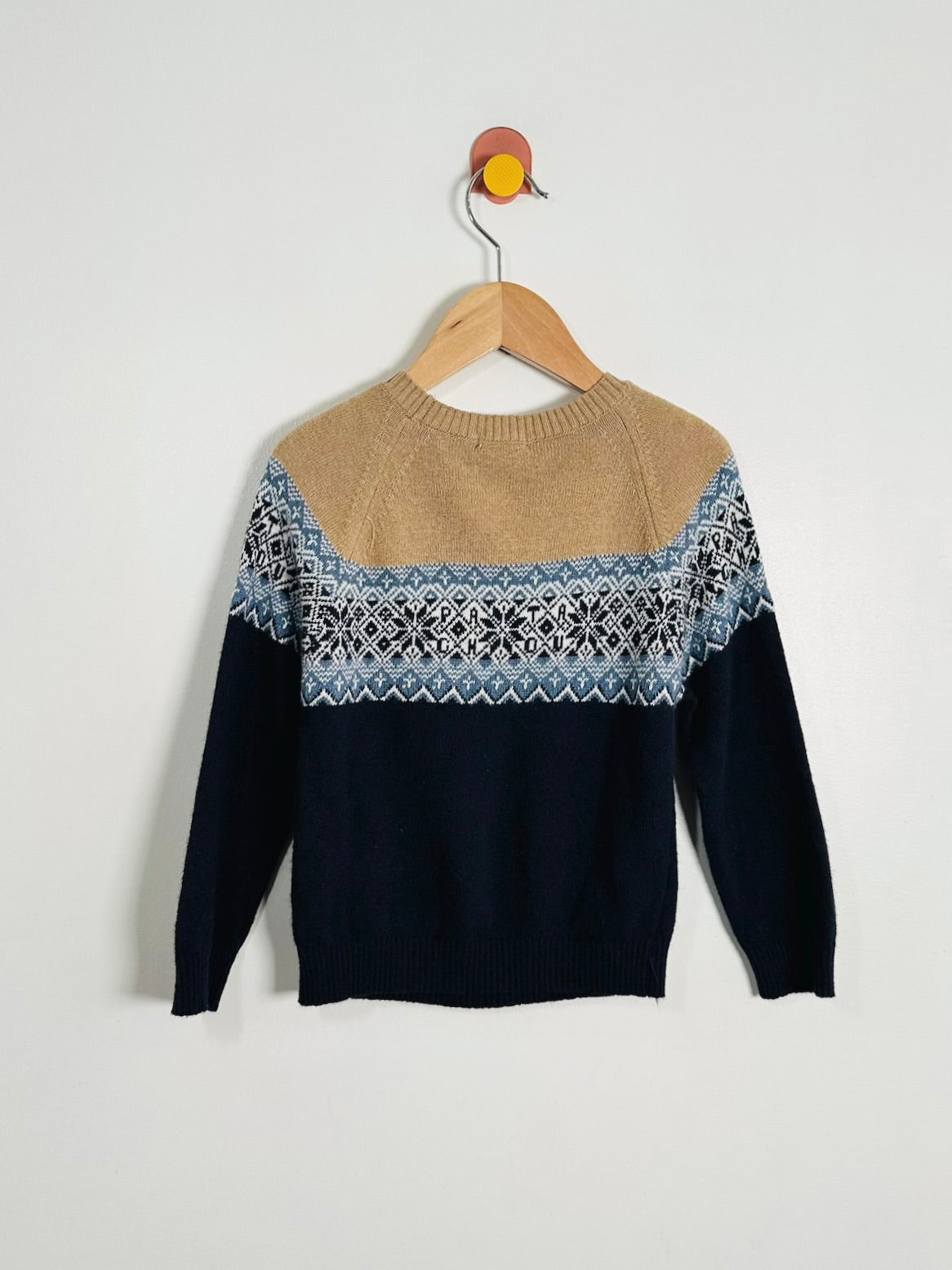 A knit crewneck sweater hanging on a wooden hanger against a plain light gray wall. The sweater has a tan yoke, a blue and white geometric Fair Isle pattern band across the chest, and dark navy sleeves and lower body.