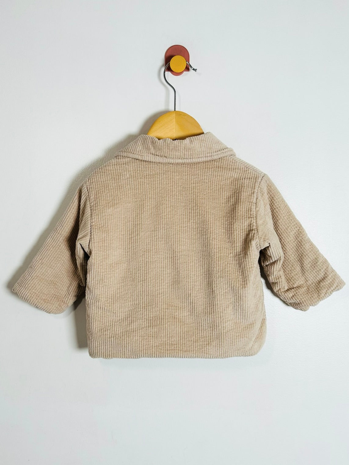A beige corduroy jacket with three-quarter sleeves, shown from the back and hanging on a wooden hanger against a plain white wall.