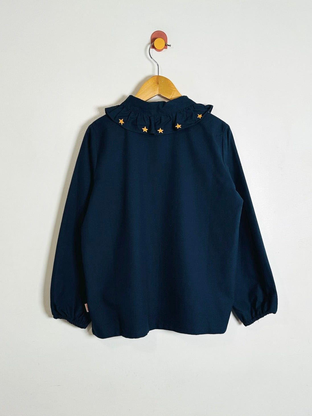 A navy blue long-sleeve button-up shirt with a ruffled Peter Pan collar decorated with small gold star appliqués, hanging on a wooden hanger against a plain white background.