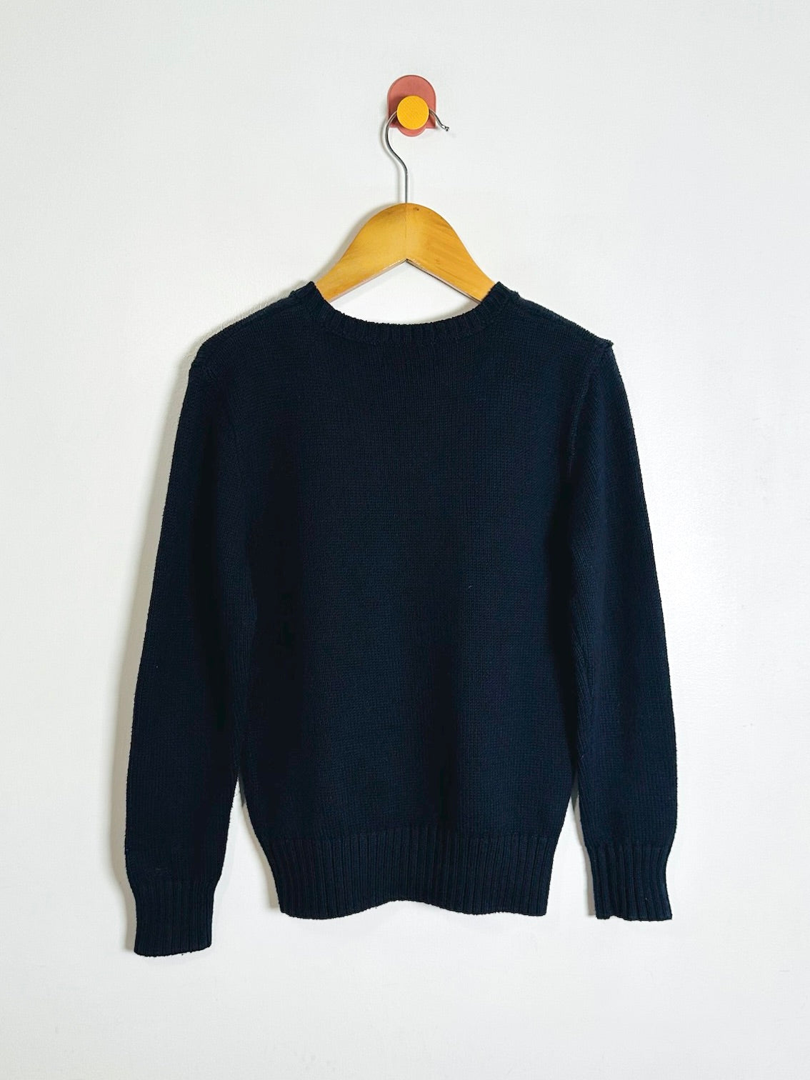 A dark navy blue knit sweater hanging on a wooden hanger against a plain white wall.
