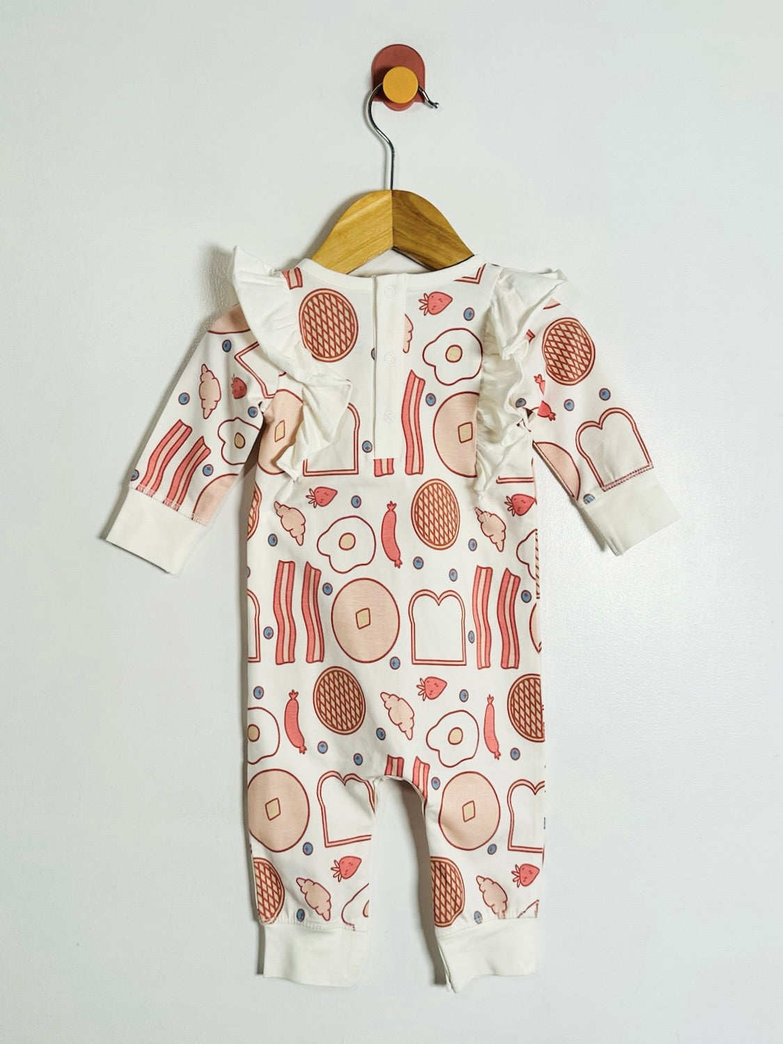 A baby long-sleeve footless romper hanging on a wooden hanger against a plain white wall. The romper is off-white with a whimsical food-themed print featuring red, orange, and brown illustrations of pancakes, bacon, eggs, toast, and fruit.