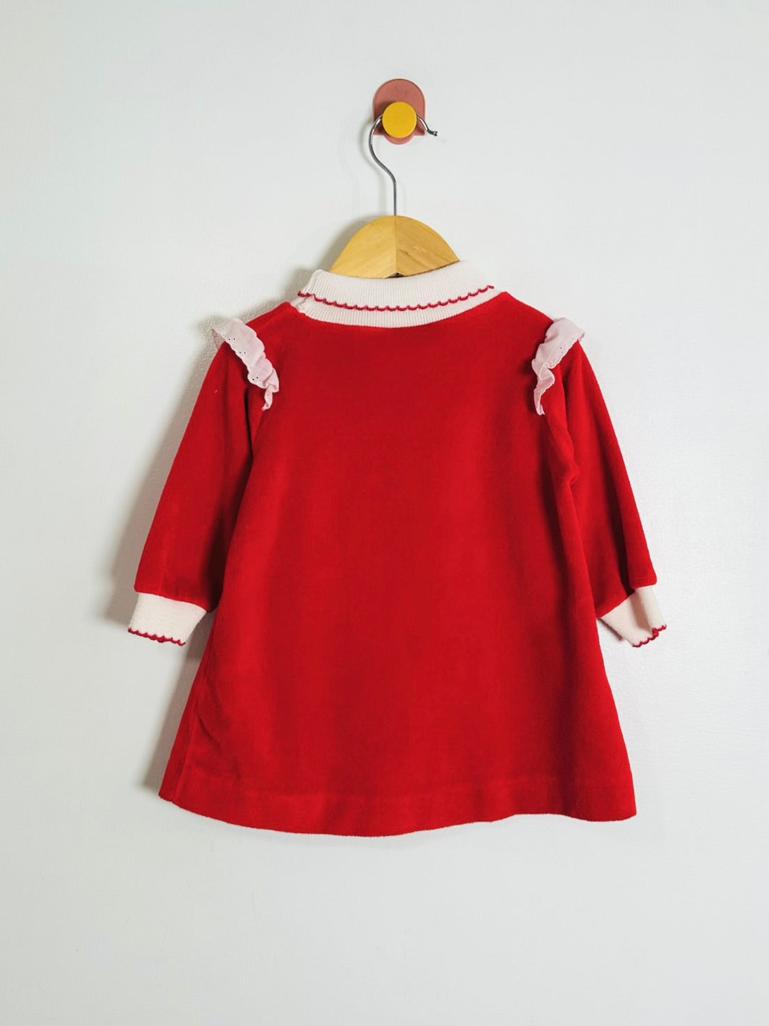 A red long-sleeve dress with white ruffle trim at the collar and cuffs, hanging on a wooden hanger against a plain white wall.