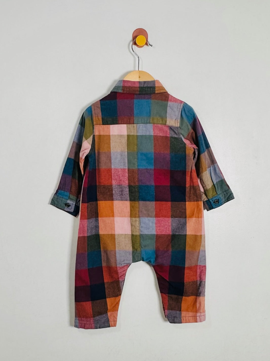 A children's long-sleeve plaid romper hanging on a wooden hanger against a plain light gray wall. The garment features a multicolor check pattern with red, blue, orange, green, pink, and brown tones.