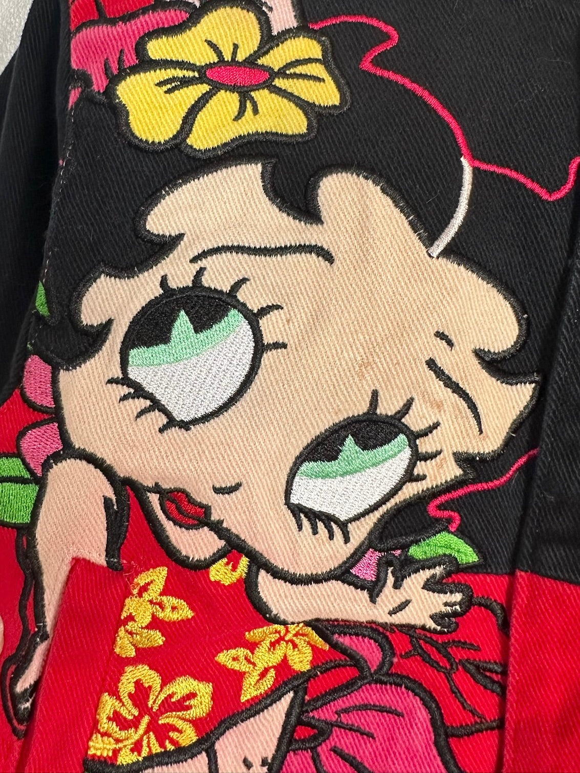 Close-up of a black jacket featuring a large embroidered cartoon Betty Boop face and floral details in red, yellow, green, and pink.