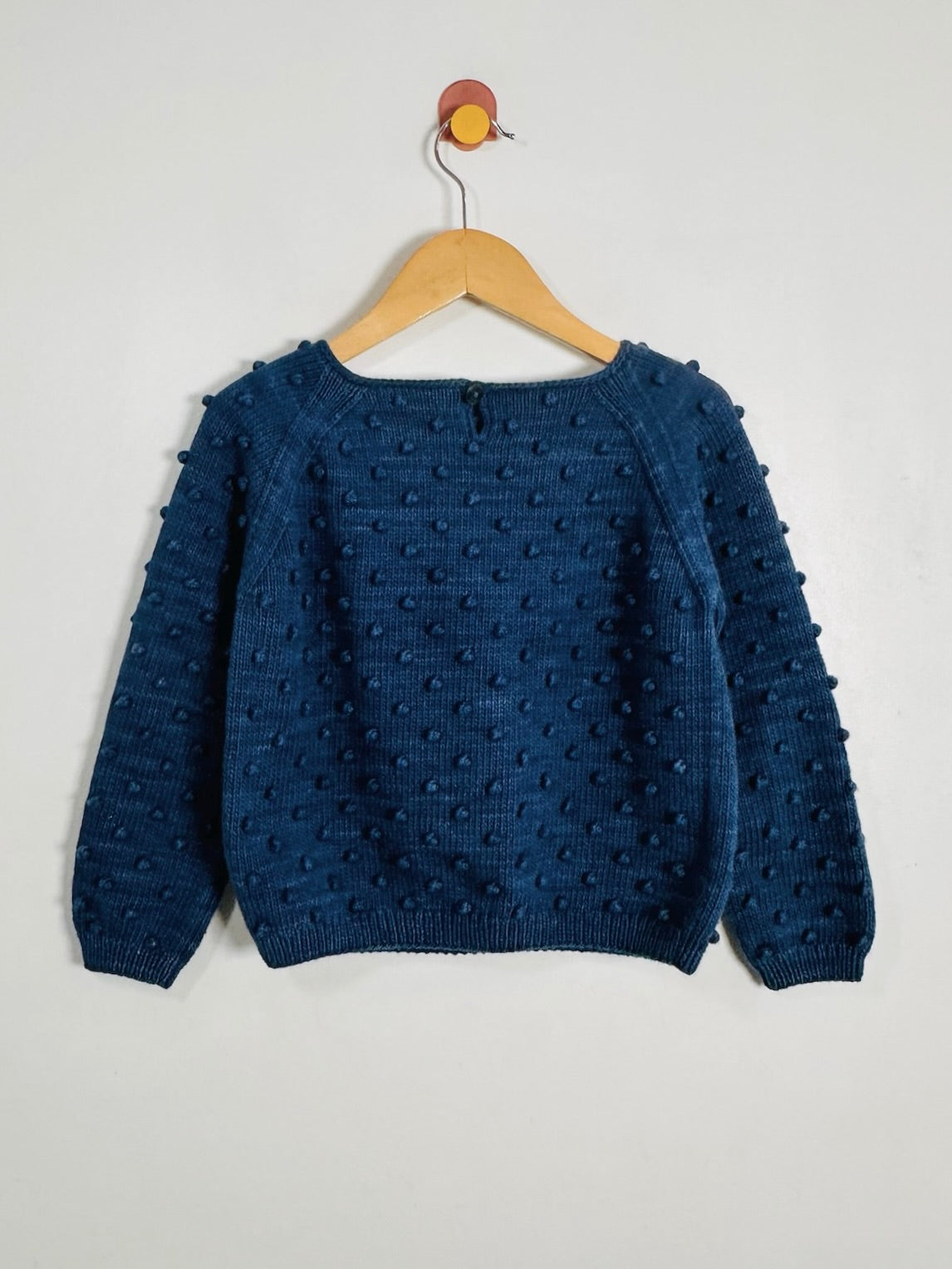 A dark blue textured knit sweater with raised pom-pom details, hanging on a wooden hanger against a plain off-white wall.