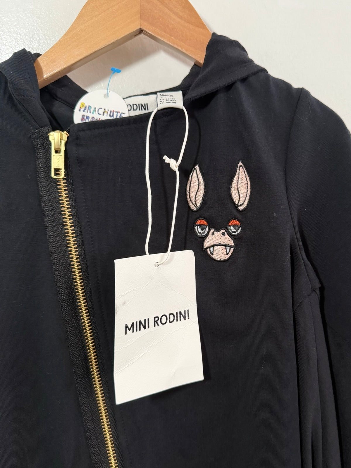 A black zip-up hoodie from Mini Rodini hanging on a wooden hanger against a white wall. The hoodie features a gold zipper, a white hangtag, and an embroidered patch of a rabbit face on the chest.