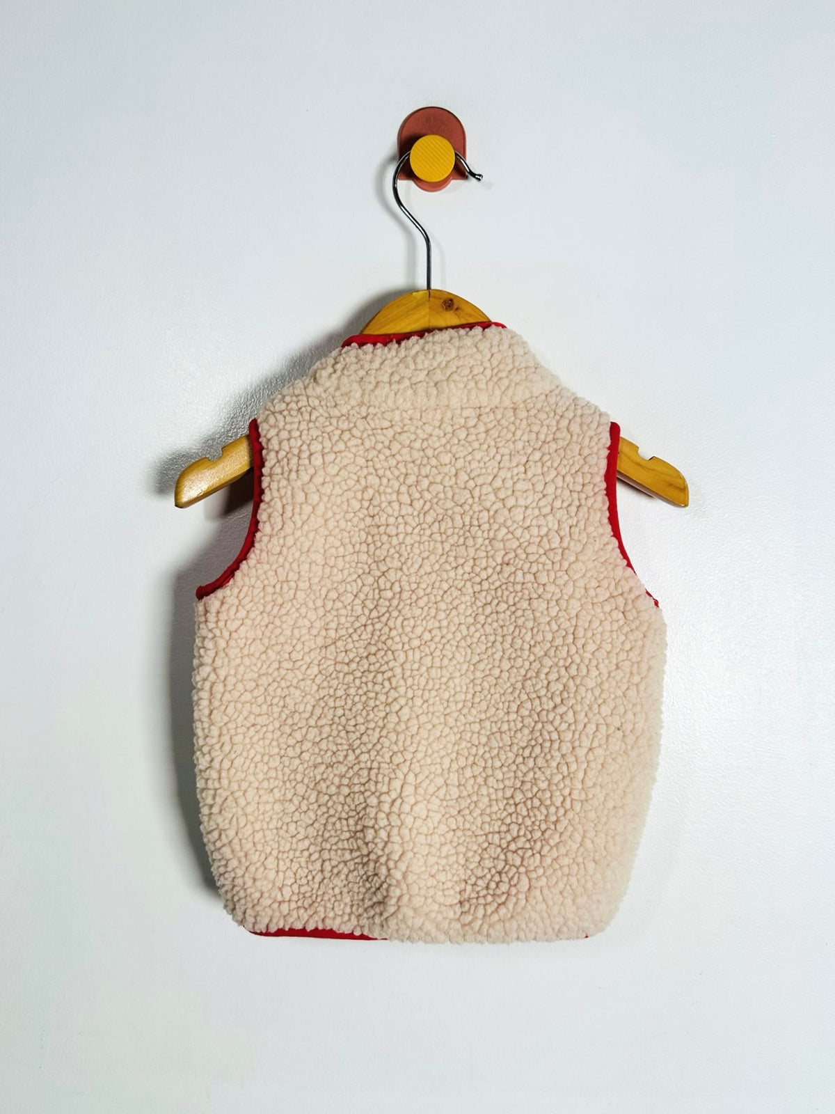 A cream-colored sherpa fleece vest with red trim, hanging on a wooden hanger against a plain white wall.