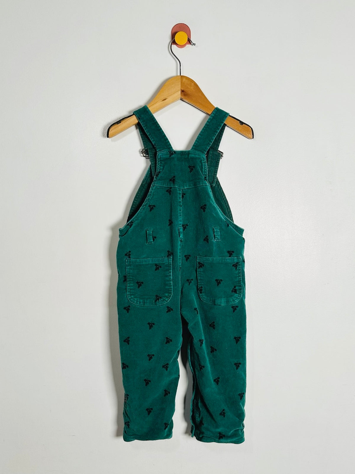 A pair of toddler green corduroy bib overalls with a small black embroidered pattern, hanging on a wooden hanger against a plain white wall.