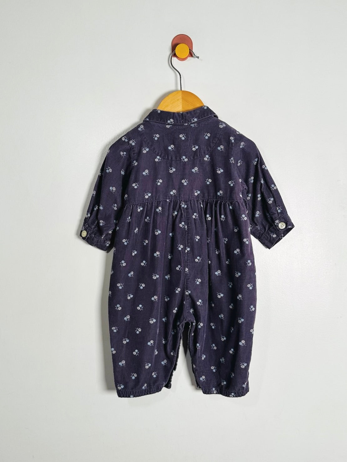 A baby romper in dark navy blue with a small white floral pattern, hanging on a wooden hanger against a plain light gray wall.
