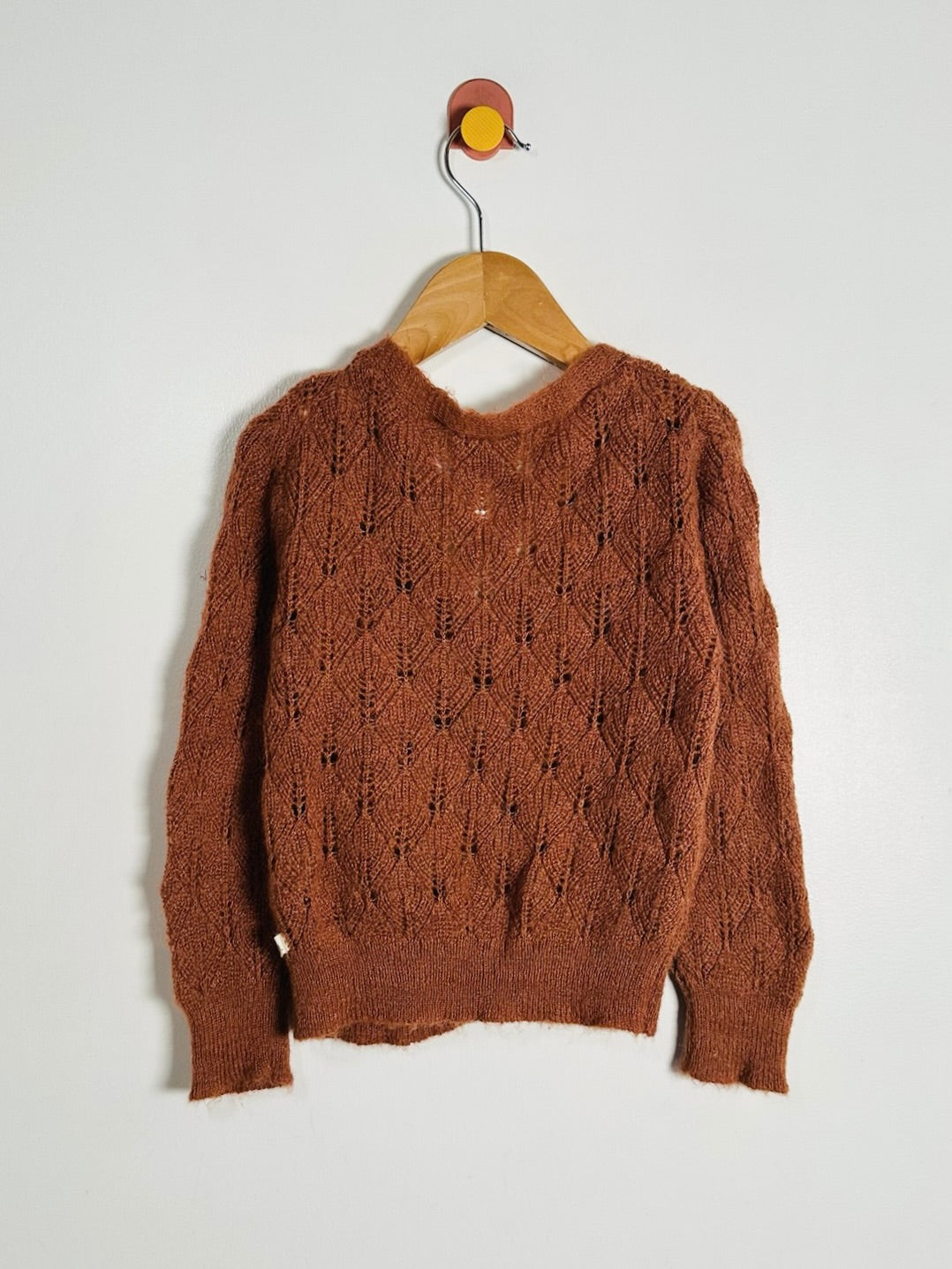 A rust-brown cable-knit sweater hanging on a wooden hanger against a plain light gray wall.
