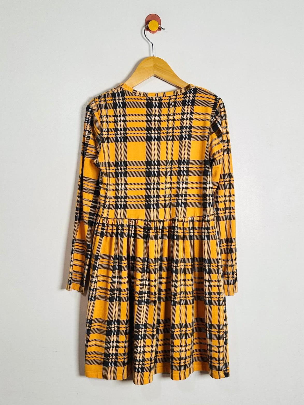 A yellow and black plaid long-sleeve dress hanging on a wooden hanger against a plain light gray wall.