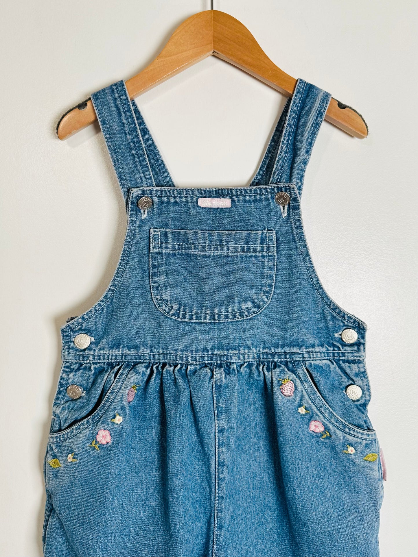 Blue denim overalls with floral embroidery on a hanger against a white background