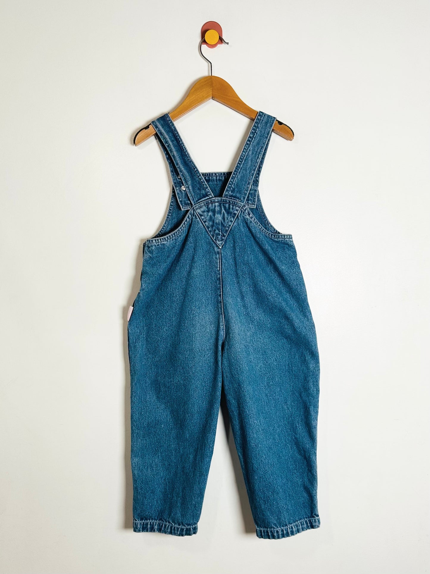 Blue denim overalls hanging on a wooden hanger against a white background