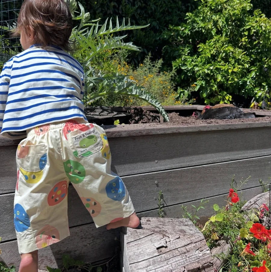 A toddler wearing cream-colored shorts with colorful painted circle patterns and a blue and white striped shirt stands beside a wooden garden planter in a sunny outdoor garden setting.
