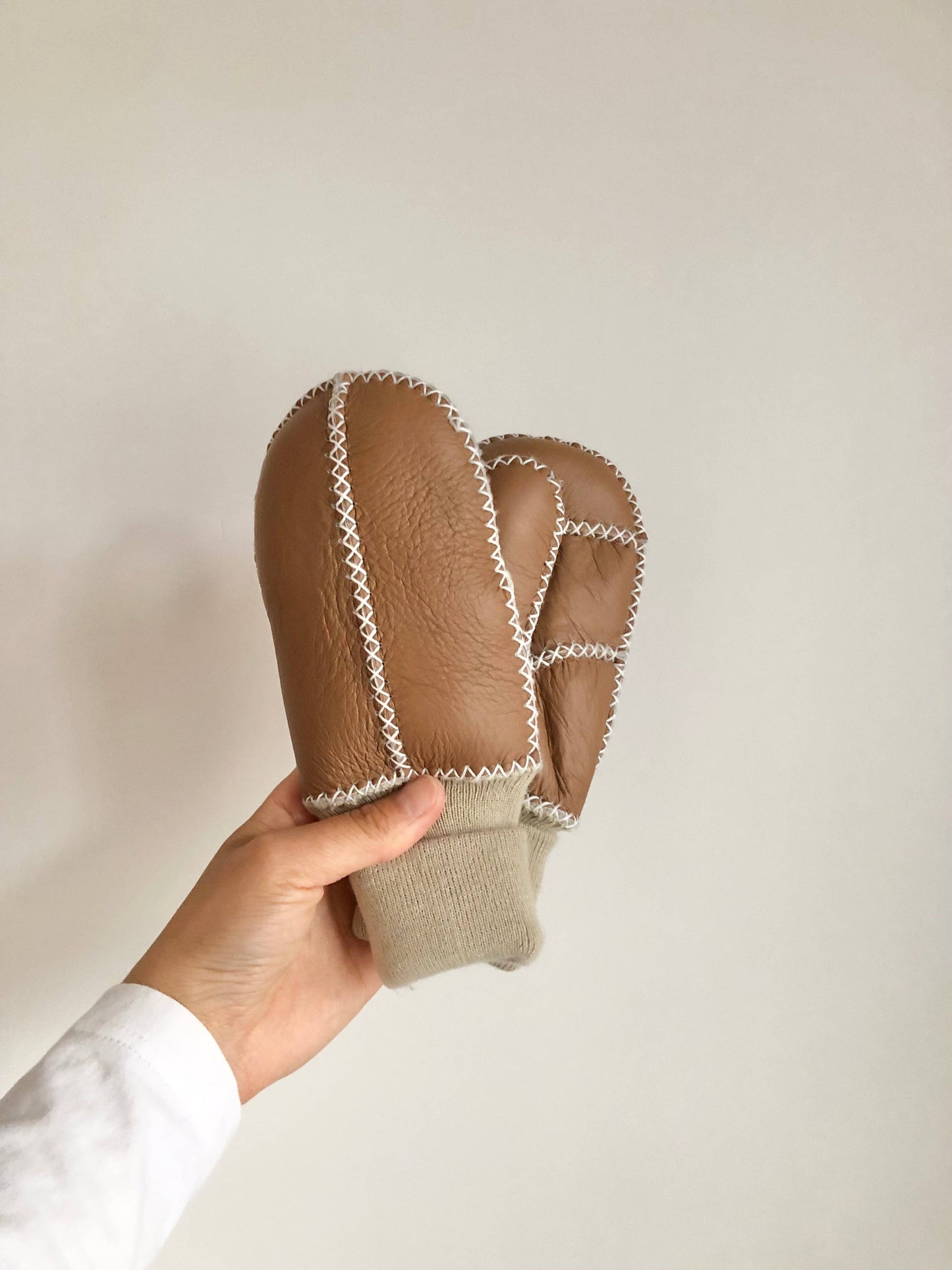 Hand holding a pair of brown leather mittens against a white background