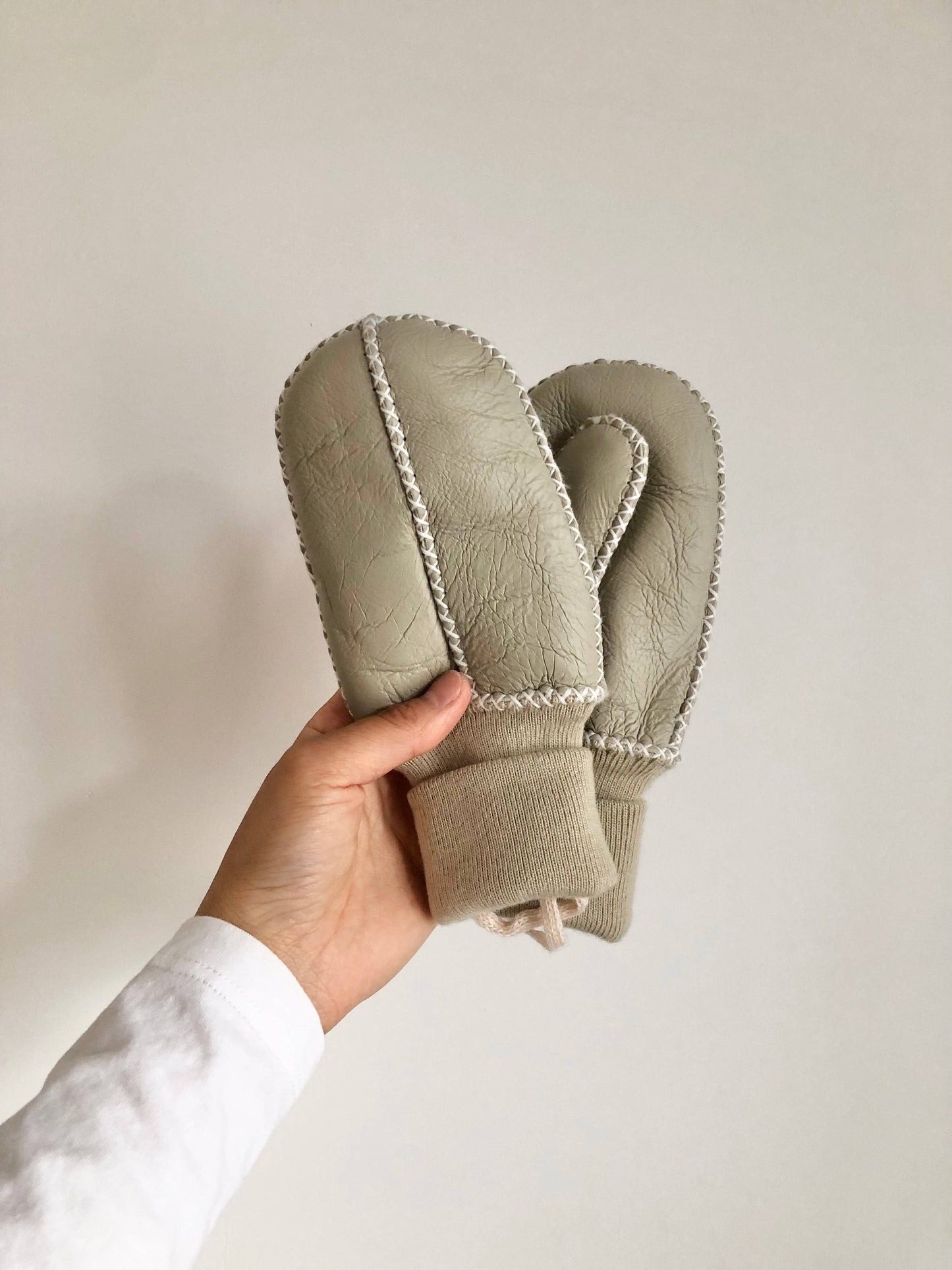 Hand holding a pair of beige mittens with white trim against a plain background