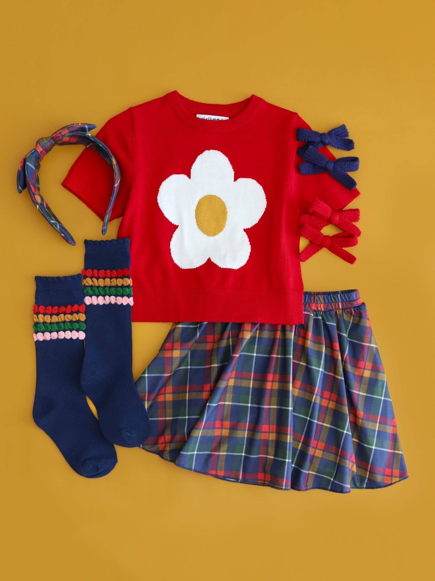Children's outfit with red shirt with flower design, plaid skirt, colorful socks, and headband on a yellow background