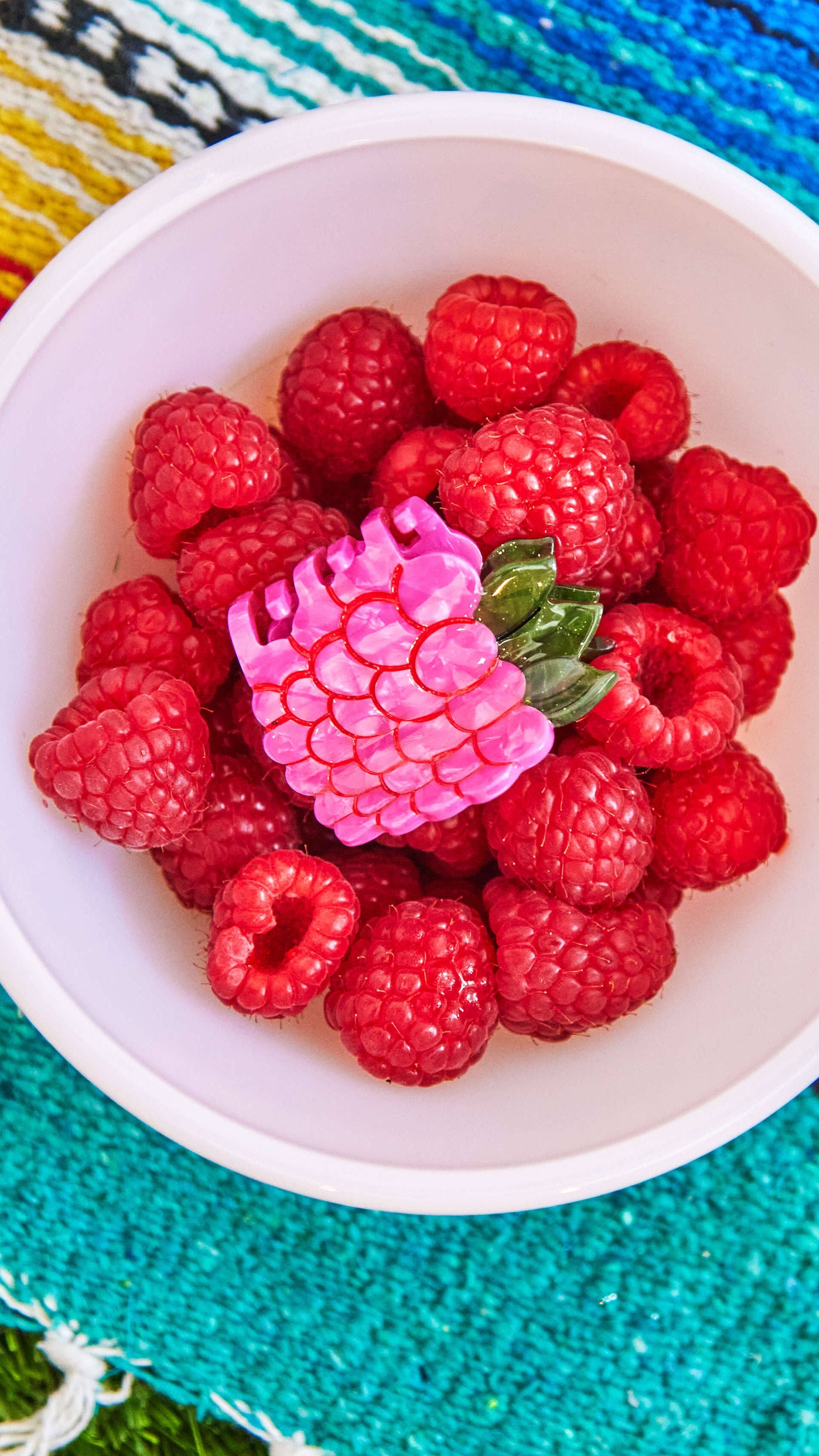 White bowl filled with red raspberries on a colorful woven mat.