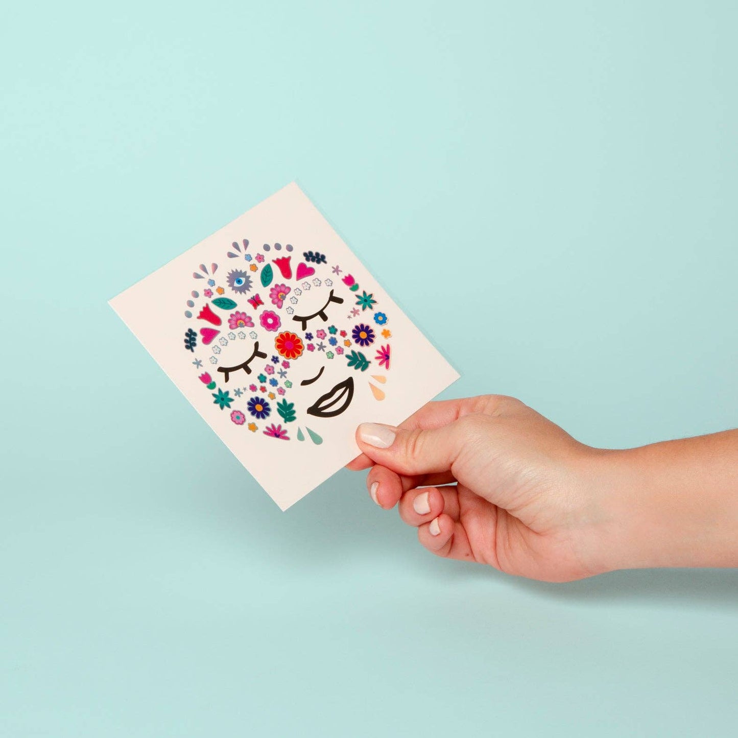 Hand holding a square card with colorful floral and face design against a light blue background