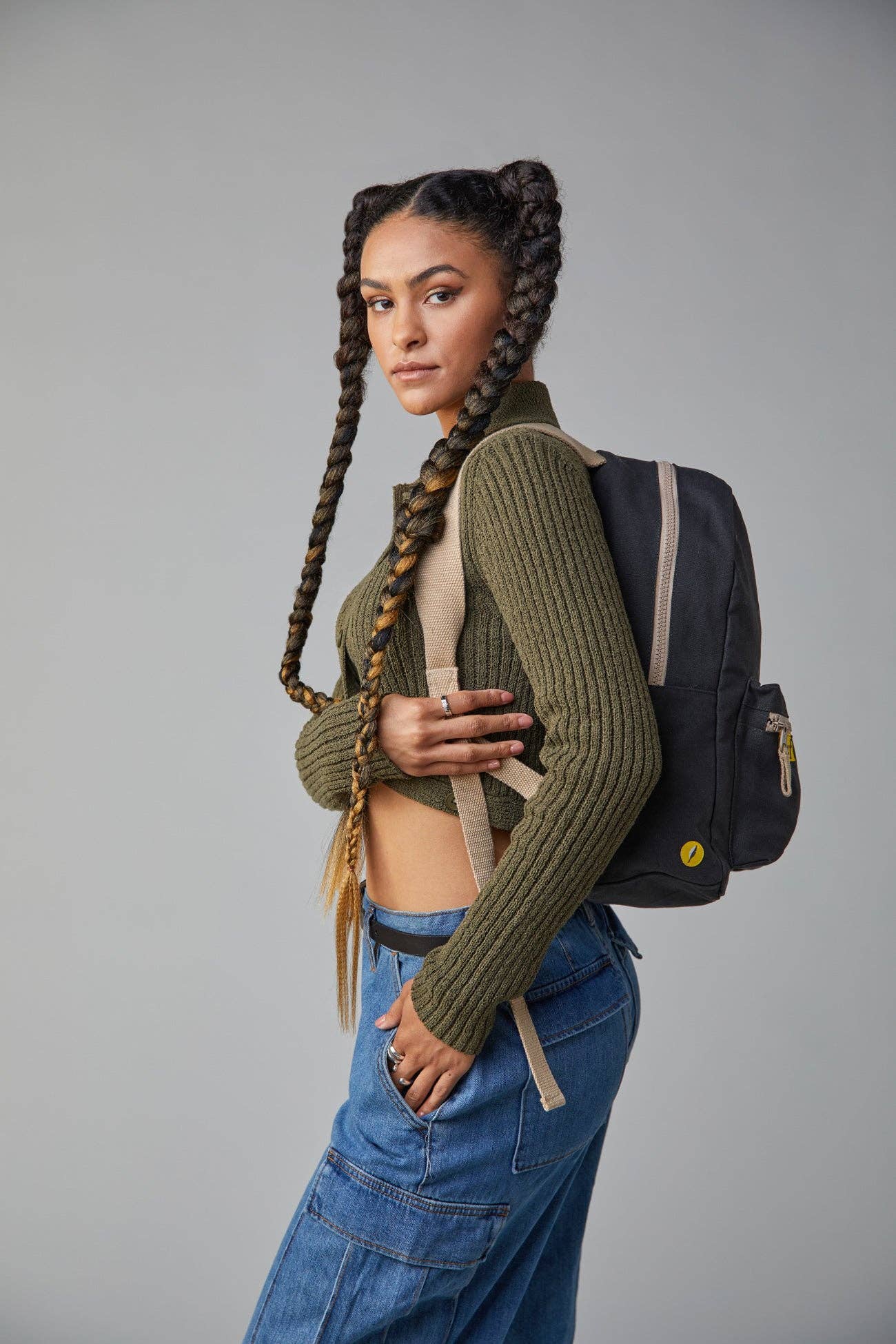 Woman with braided hair wearing a green sweater and blue jeans, standing against a gray background.