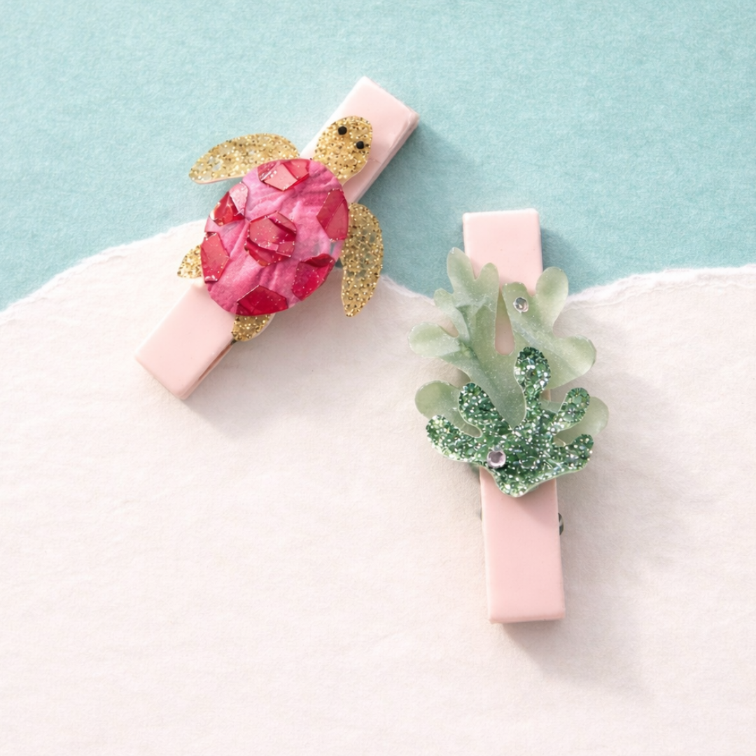Two decorative clips with a pink base, turtle design, and green leaf on a light blue and white background.