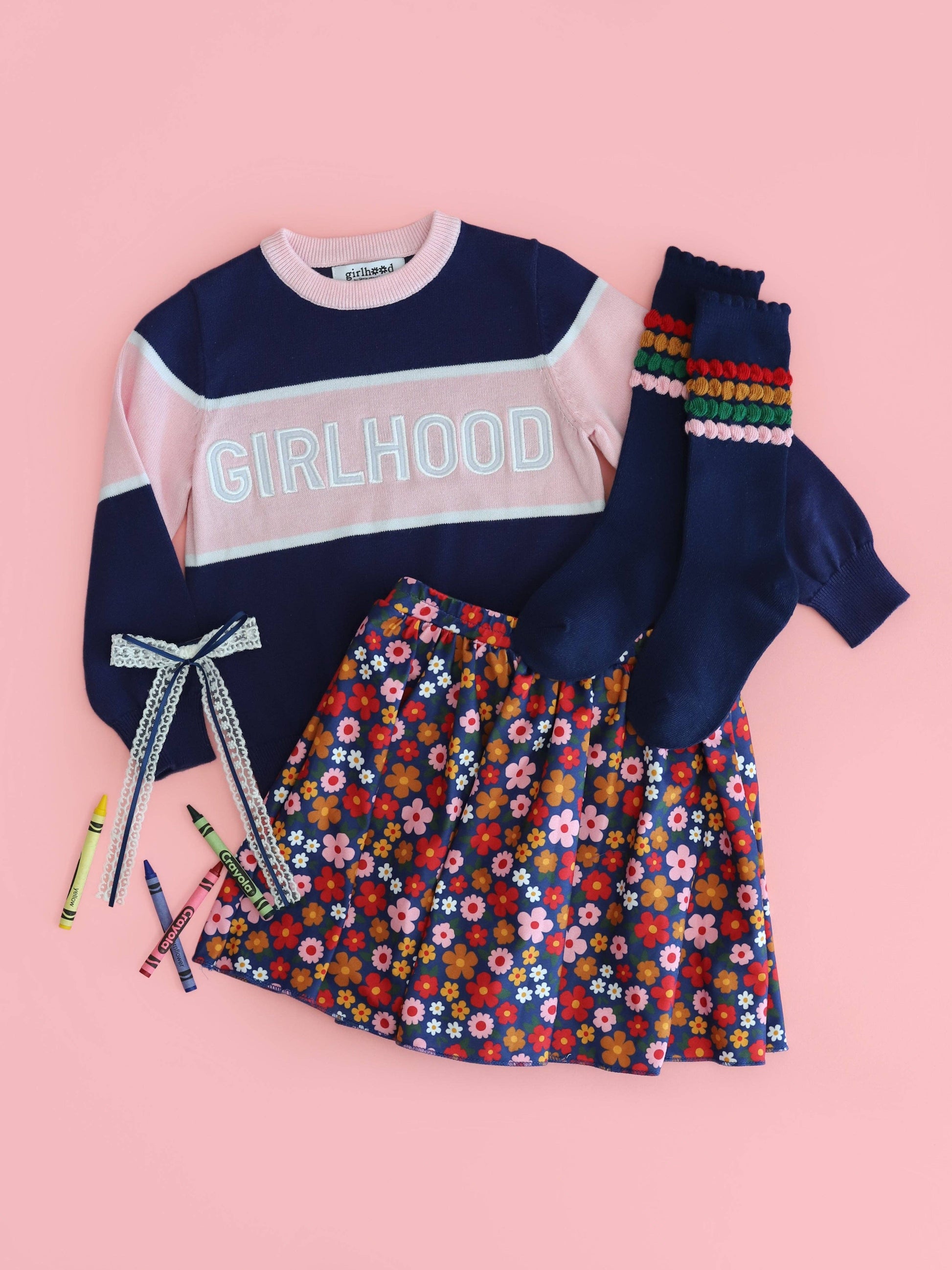 Children's outfit with navy sweater, floral skirt, and colorful arm warmers on a pink background