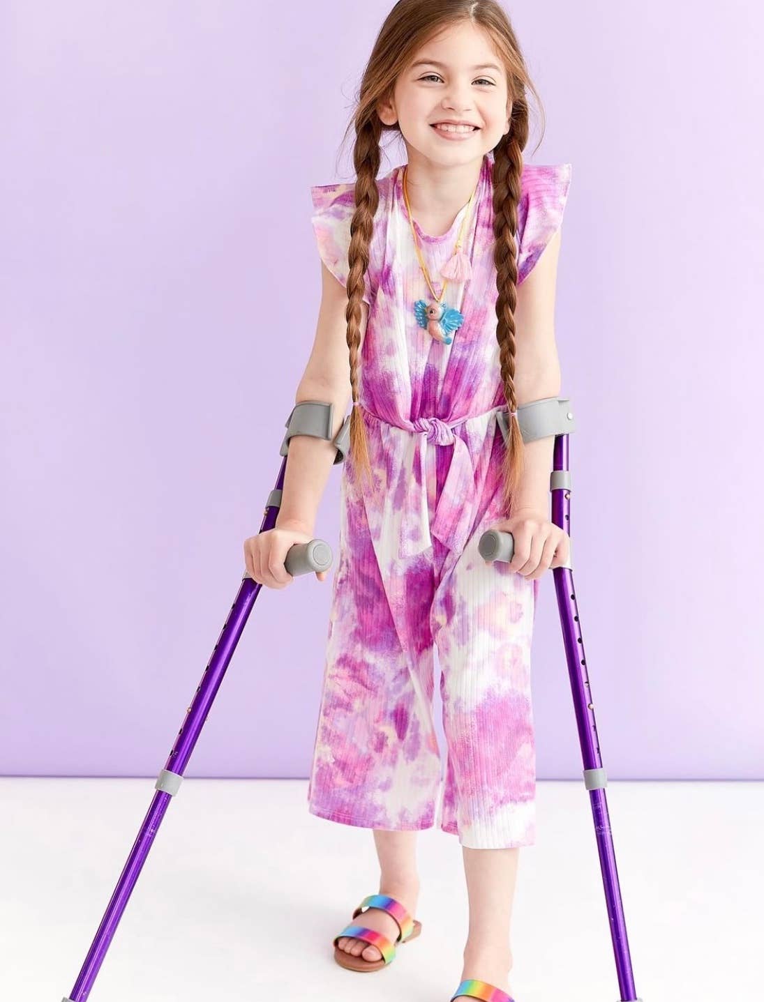 A young girl with long braided hair stands on a white floor using purple crutches, wearing a pink and white tie-dye sleeveless dress, rainbow sandals, and a butterfly necklace against a light purple background.