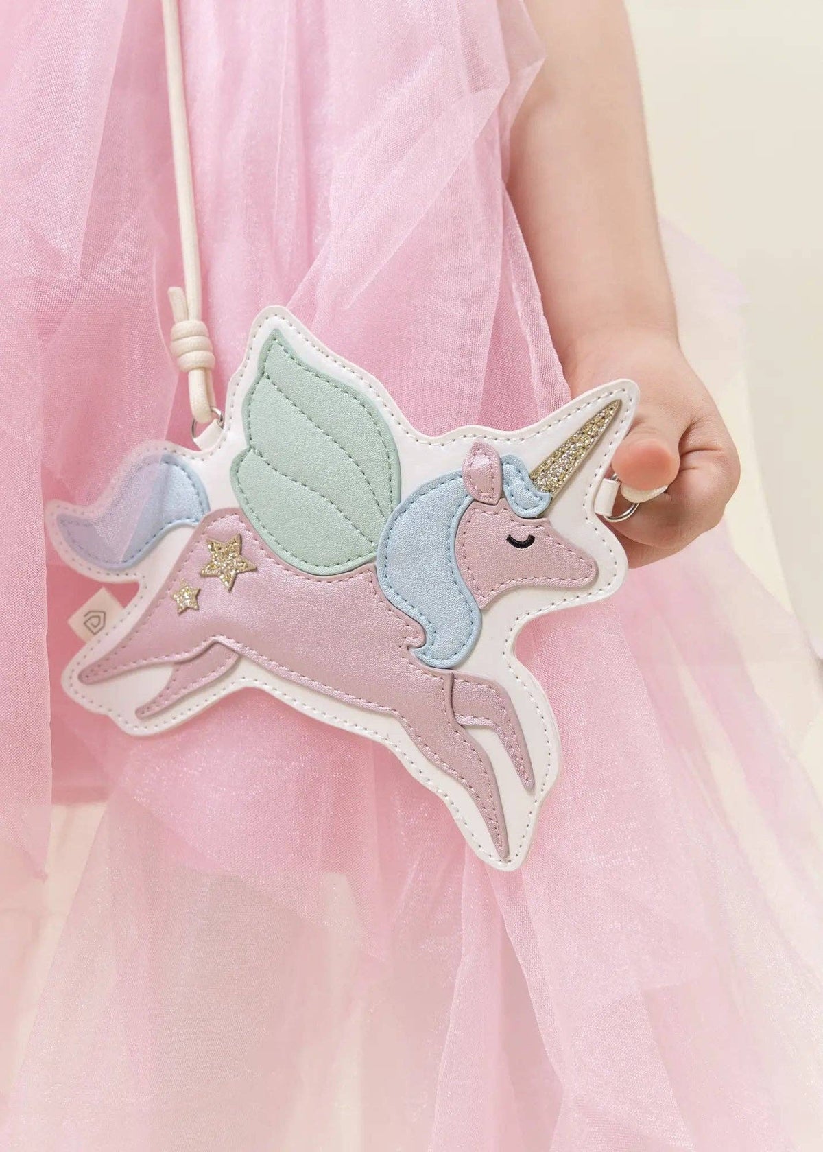 A child wearing a pink tulle dress holds a pastel unicorn-shaped crossbody bag. The bag features a pink body, mint green wings, a light blue mane, a gold horn, and embroidered stars, with a white strap.