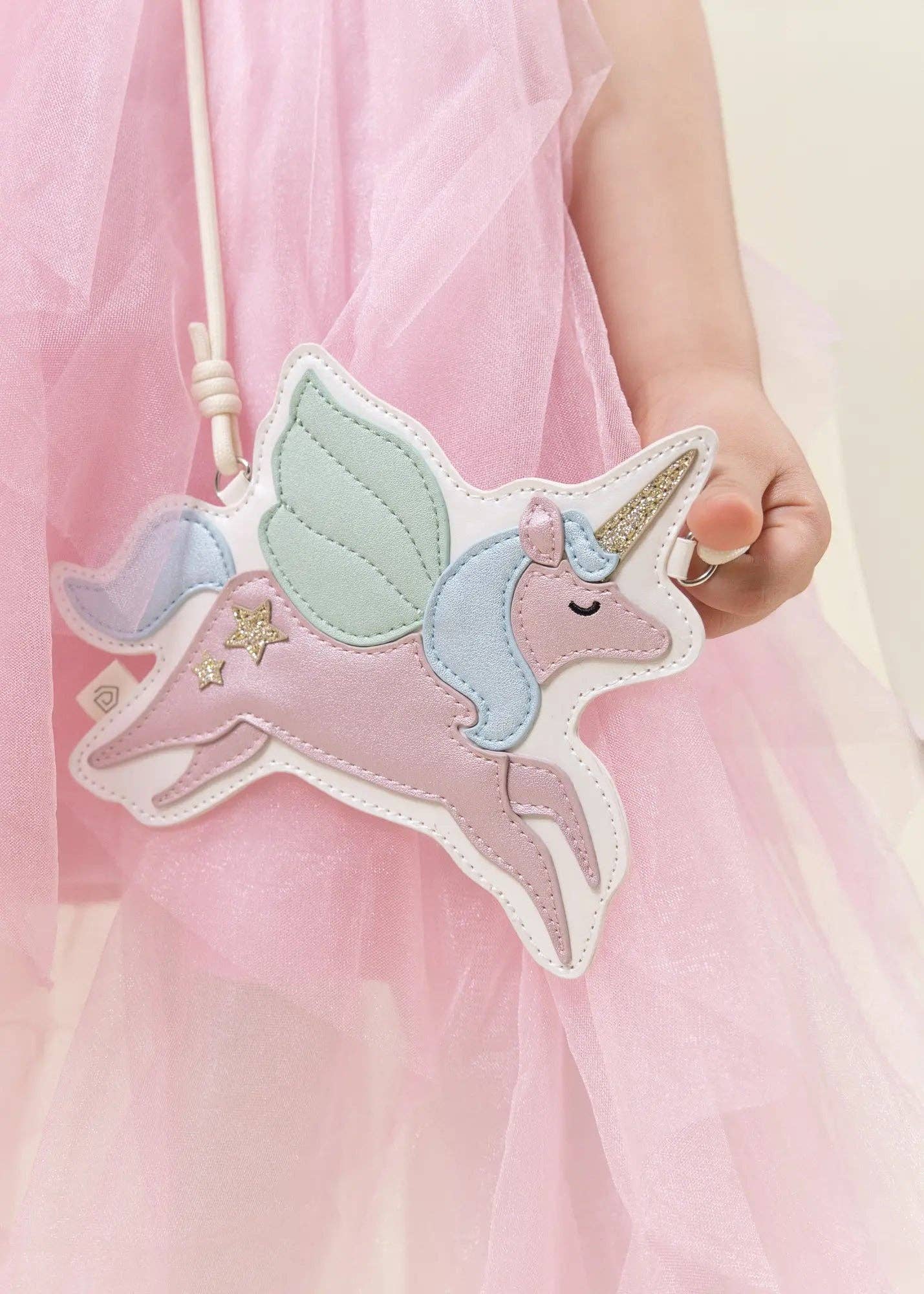 A child wearing a pink tulle dress holds a pastel unicorn-shaped crossbody bag. The bag features a pink body, mint green wings, a light blue mane, a gold horn, and embroidered stars, with a white strap.