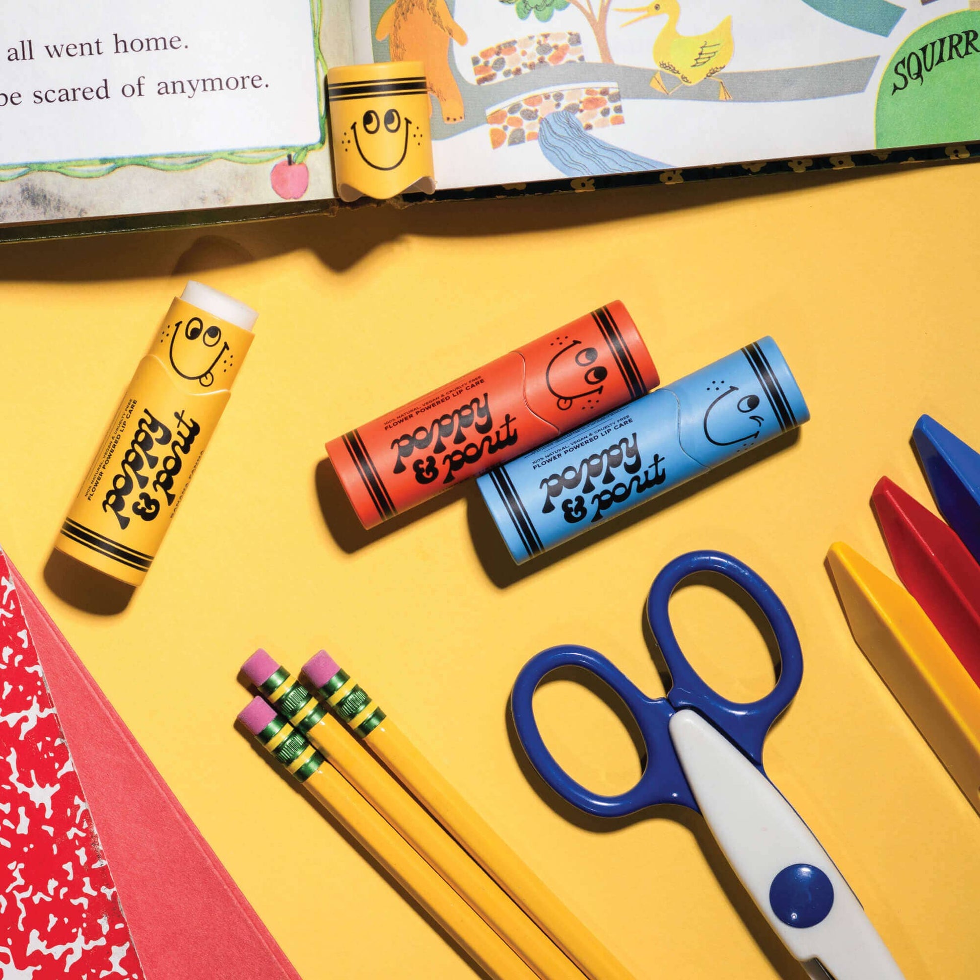 Colorful lip balms, scissors, and pencils on a yellow surface with a book in the background.