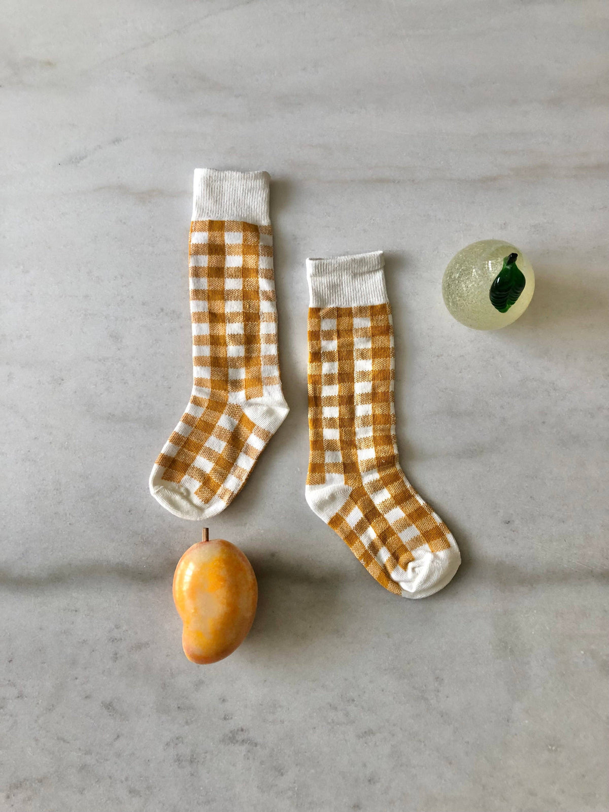 A pair of white socks with a mustard yellow gingham check pattern, styled in a flat-lay arrangement on a light gray marble surface with a small yellow fruit and a decorative glass object.
