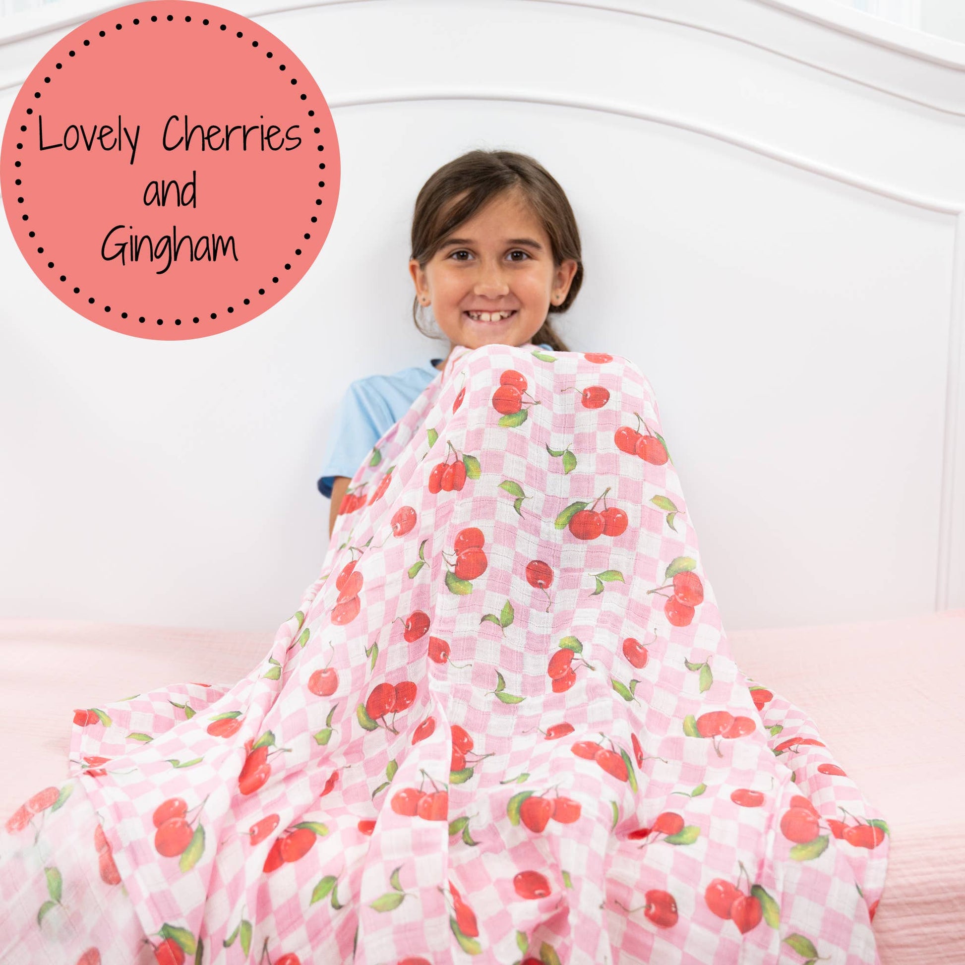Child wrapped in a pink blanket with cherry pattern, sitting on a white bed.