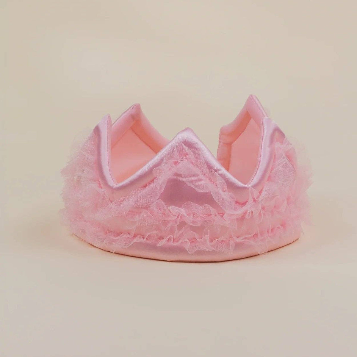 A pink ruffled tulle crown with a satin inner band, photographed on a plain beige studio background.