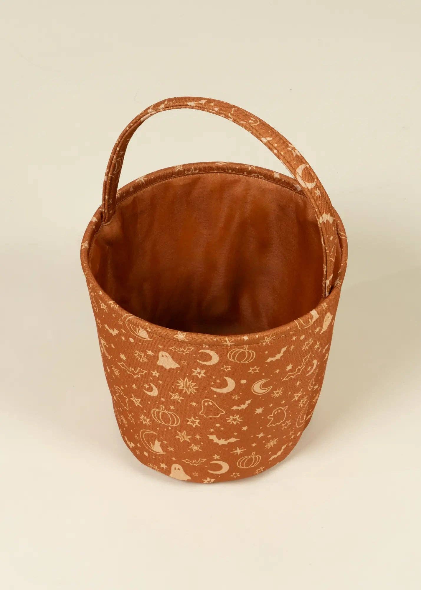 A terracotta brown fabric bucket with a single top handle, decorated with a light cream print of pumpkins, ghosts, stars, and crescent moons, set against a plain off-white background.