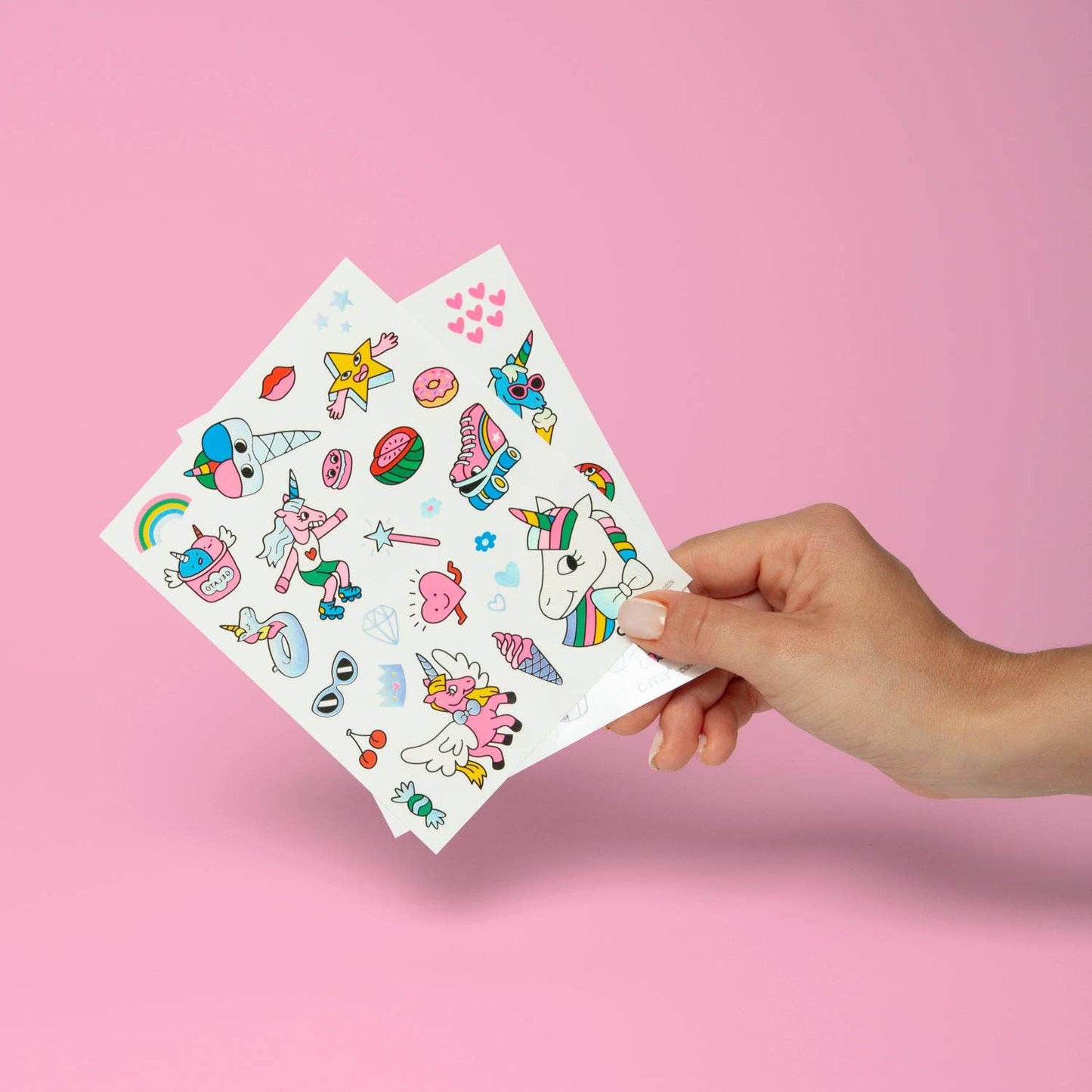 Hand holding a sheet of colorful temporary tattoos against a pink background