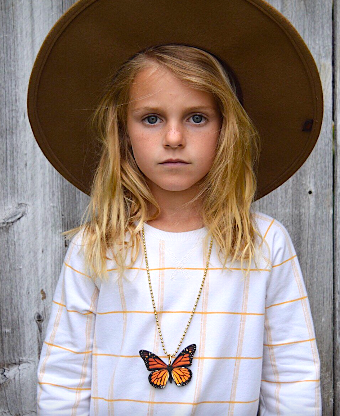 Young girl wearing a brown hat and white shirt with a butterfly necklace, standing against a wooden background.
