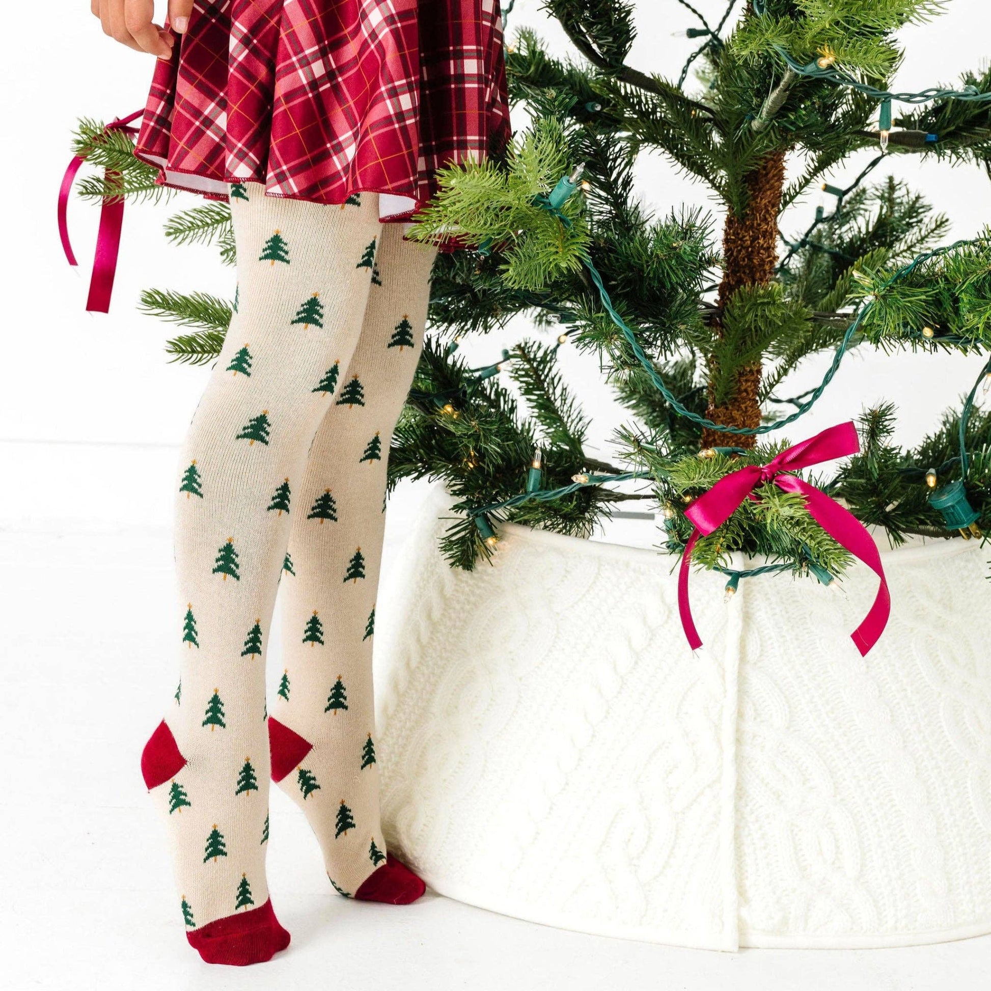 Person wearing tree-patterned tights standing next to a decorated Christmas tree.