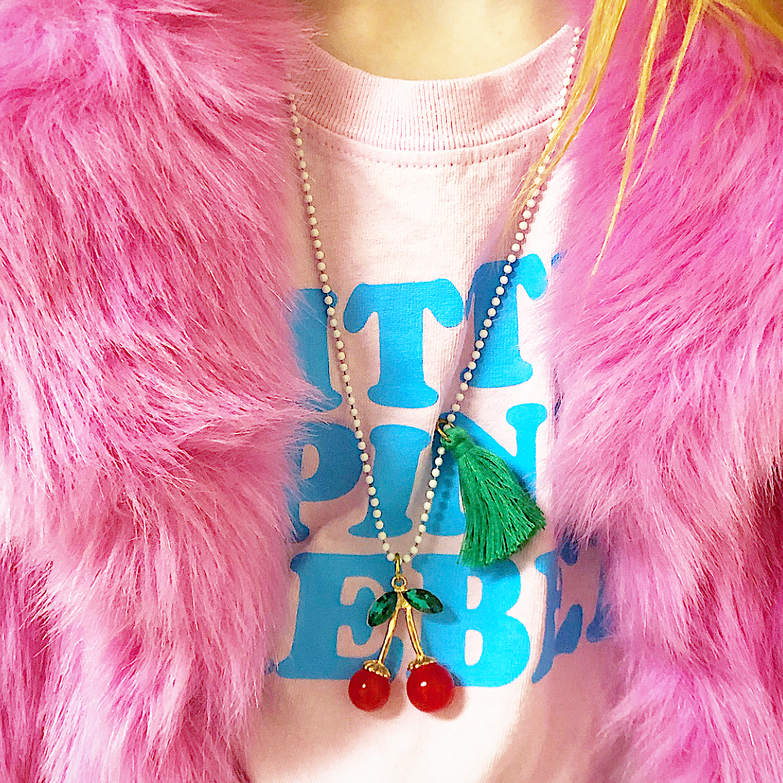 Person wearing a pink fluffy garment, blue shirt with text, and a necklace with cherry pendants.