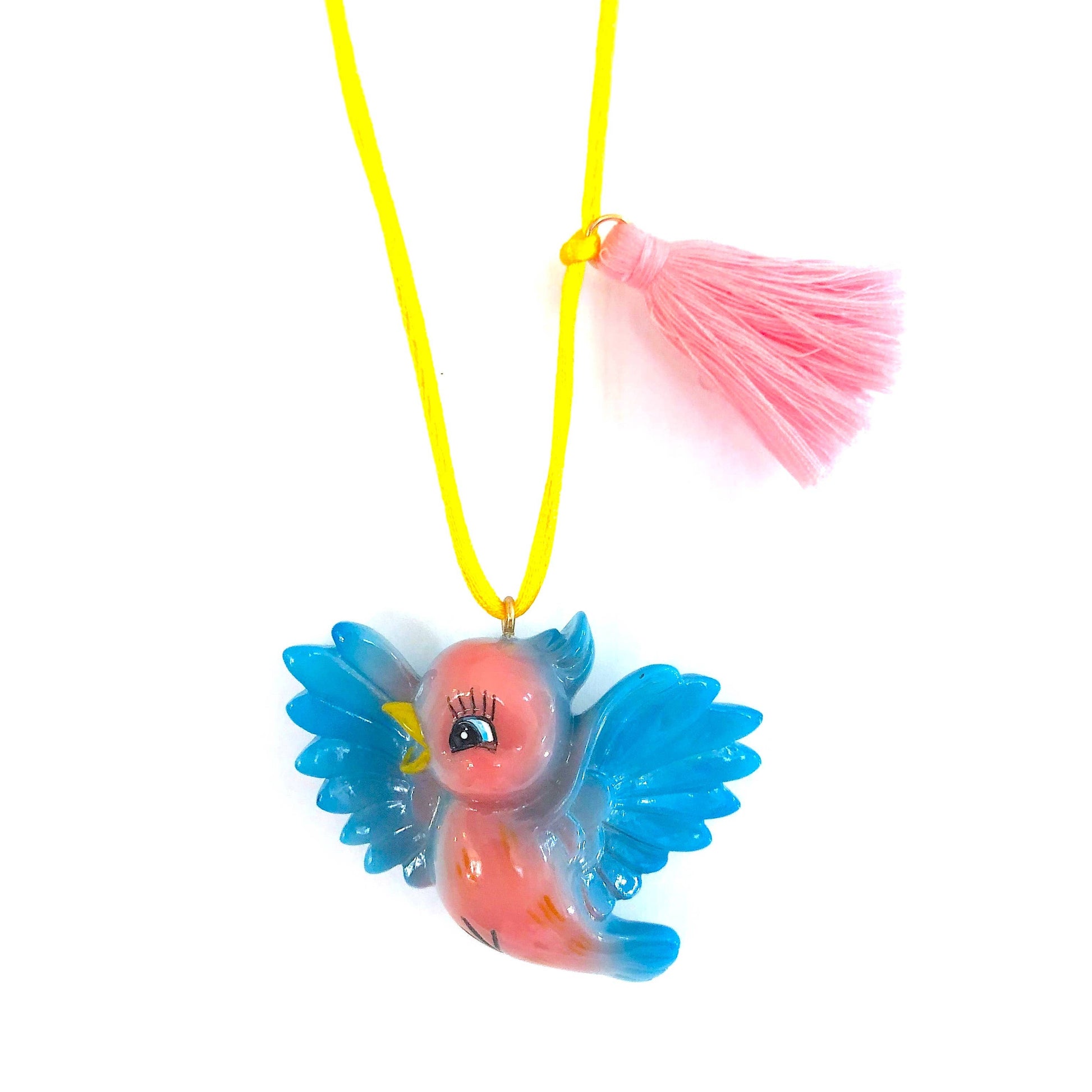 A colorful pendant necklace featuring a small bird charm with blue wings and a pink body, suspended on a bright yellow cord with a pink tassel, set against a plain white background.