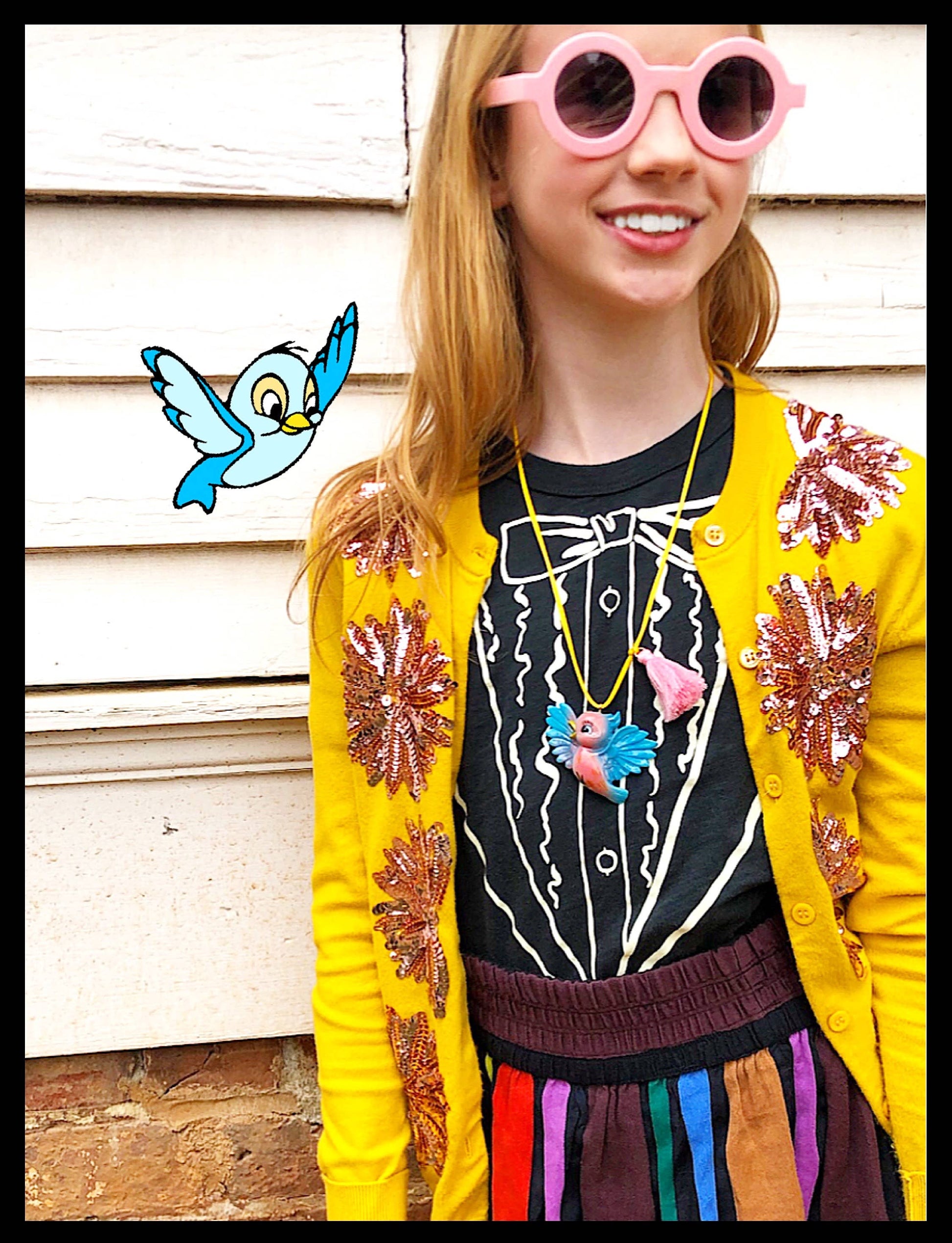 A young girl wearing a bright yellow cardigan with sequined floral patterns, a black tuxedo-print top, and colorful sunglasses, standing against a white wooden wall.
