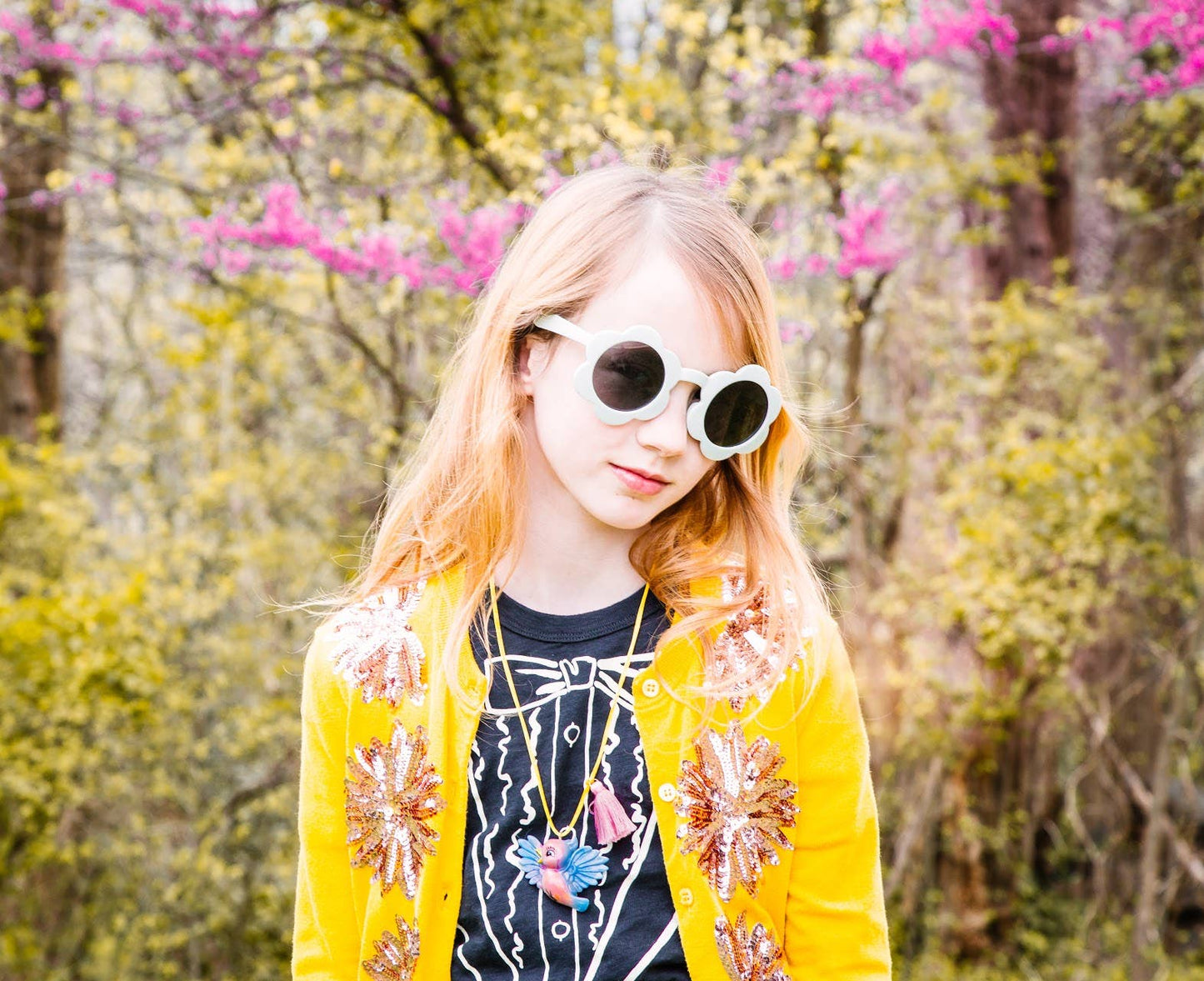 A young girl with long blonde hair wearing round white-framed sunglasses, a bright yellow embroidered cardigan over a black graphic top, and layered necklaces, standing outdoors in front of a blurred background of pink flowers and green foliage.