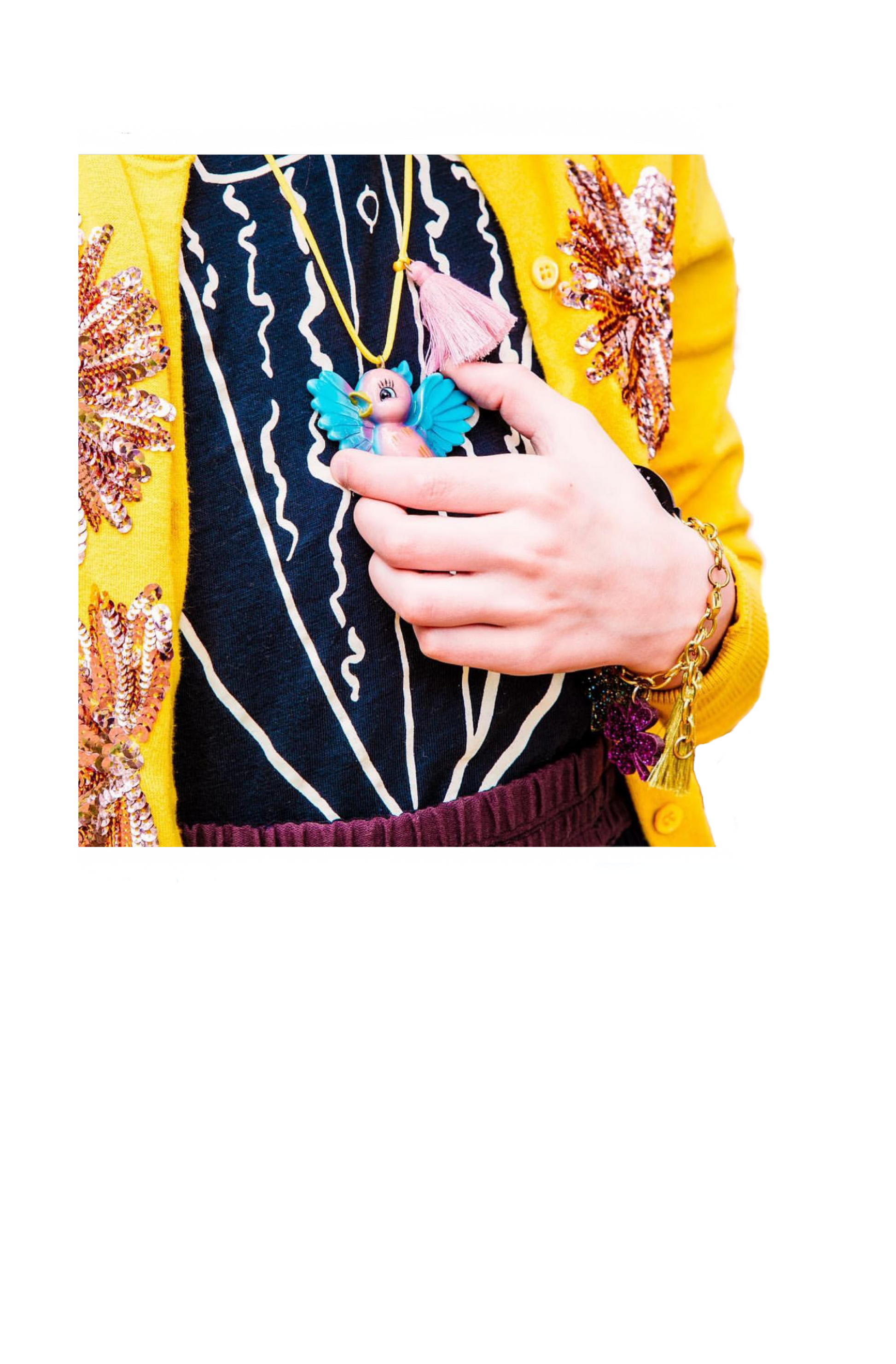 A close-up of a person holding a colorful bird-shaped pendant necklace. The person is wearing a bright yellow jacket with sequined floral details, a black and navy patterned sweater, and a purple skirt, with gold bracelets on the wrist.